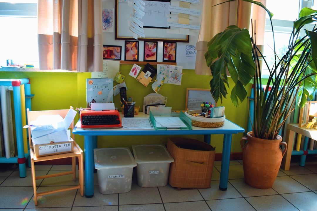 Angolo della classe con un tavolo blu, fogli di carta, contenitori e piante in vaso vicino a una finestra soleggiata.