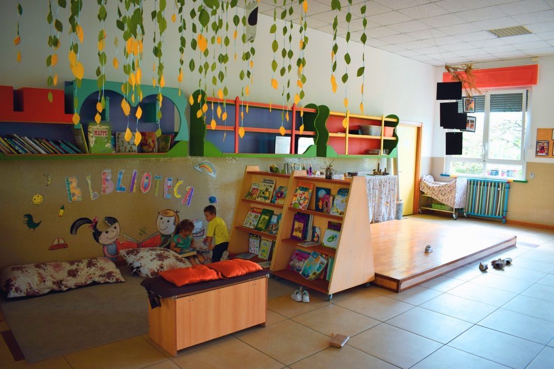 Un'area giochi colorata in classe con giocattoli, scaffali, decorazioni da appendere e un piccolo palcoscenico vicino alla finestra.
