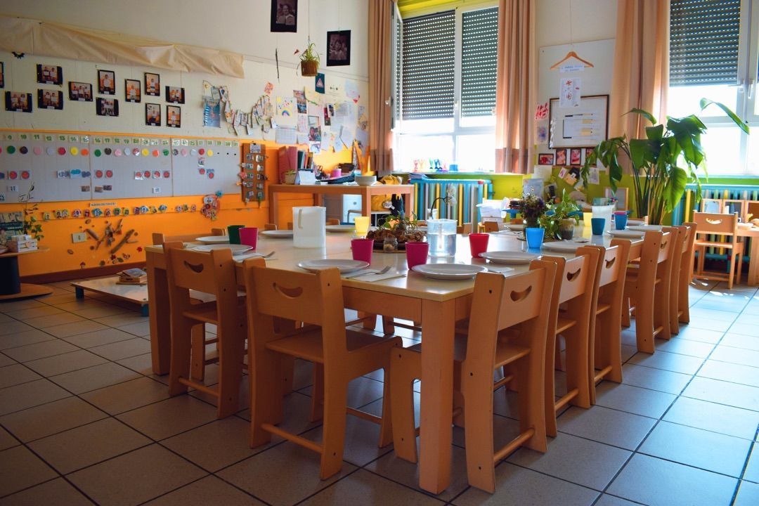 Aula per l'infanzia con tavoli e sedie in legno e pareti colorate, illuminata dalla luce del sole proveniente dalle finestre.