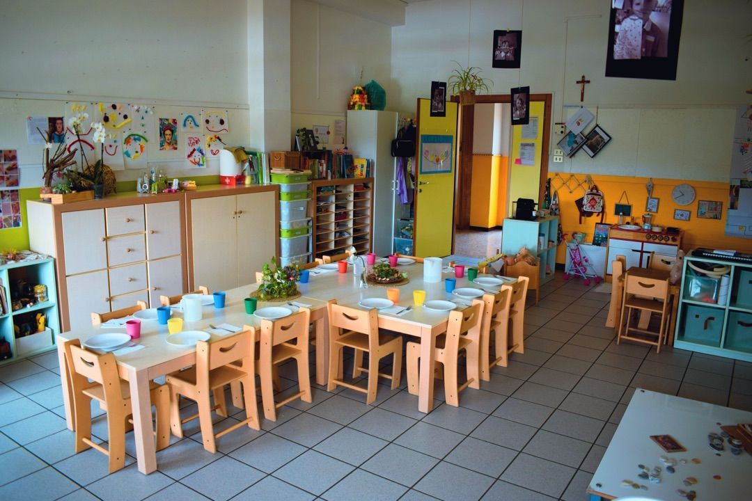 Aula per bambini con tavolini di legno, sedie, armadietti e materiali didattici colorati.