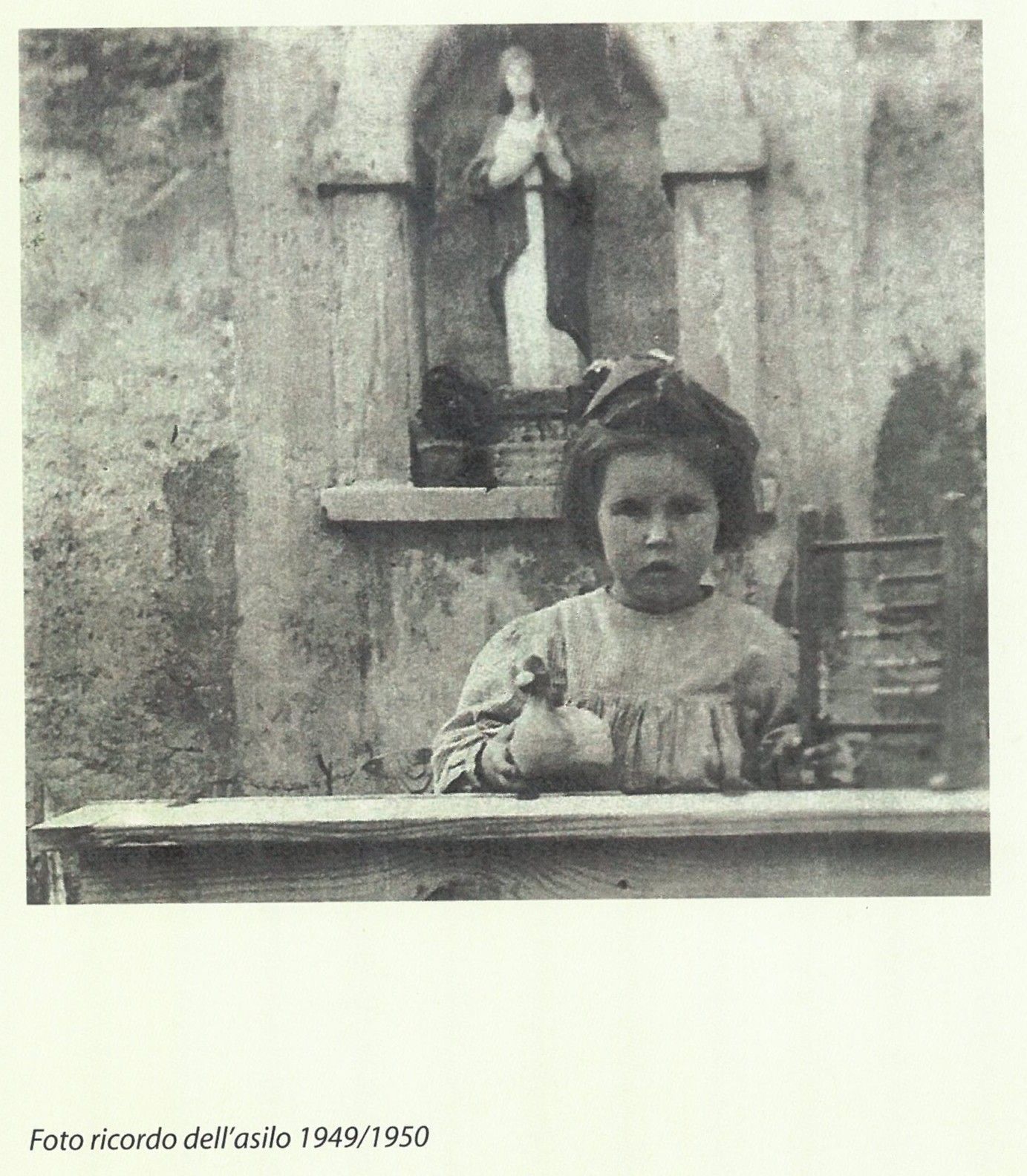 Un bambino in piedi dietro un tavolo di fronte a una nicchia nel muro con una piccola statua sopra.