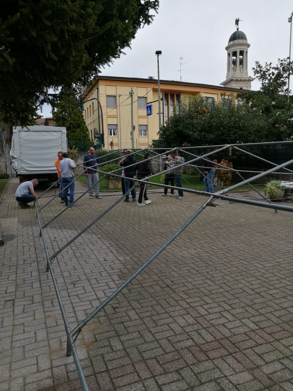 Alcune persone stanno erigendo una barricata metallica in una piazza lastricata di ciottoli, vicino a un edificio giallo e al campanile di una chiesa.
