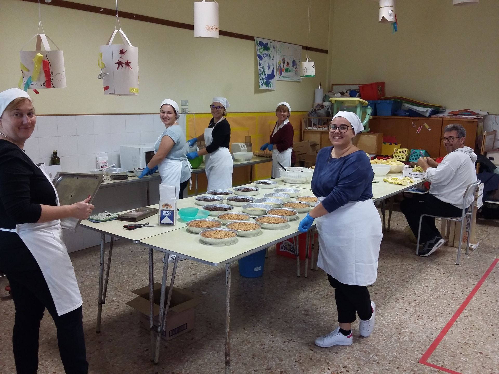 In una luminosa cucina comune, alcune persone con il grembiule preparano il cibo ai tavoli.