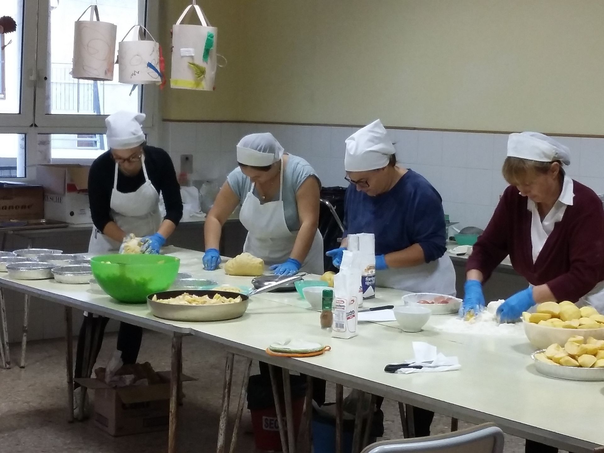 Persone con grembiuli e retine per capelli preparano il cibo insieme attorno a un lungo tavolo in cucina.