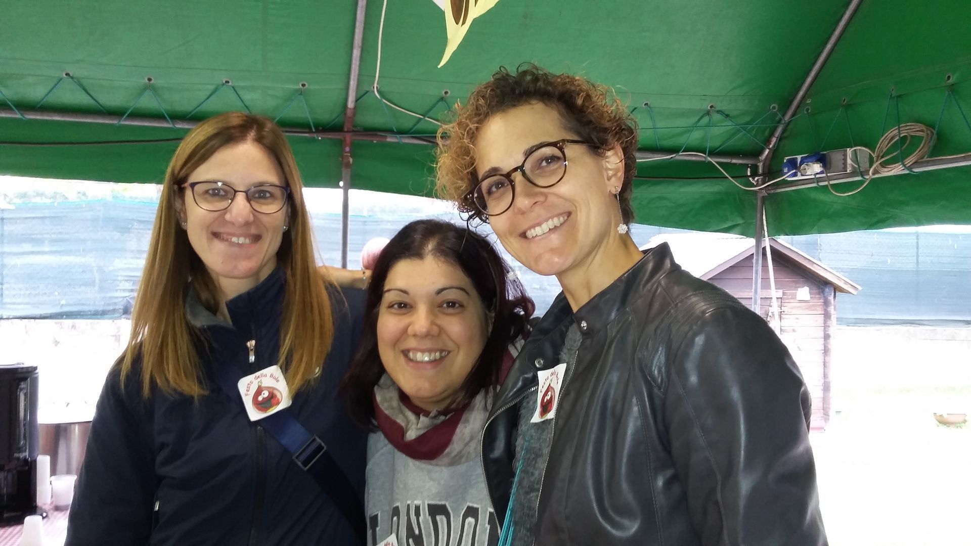 Tre persone sorridenti sotto una tettoia verde, in piedi vicine l'una all'altra in un ambiente chiuso.