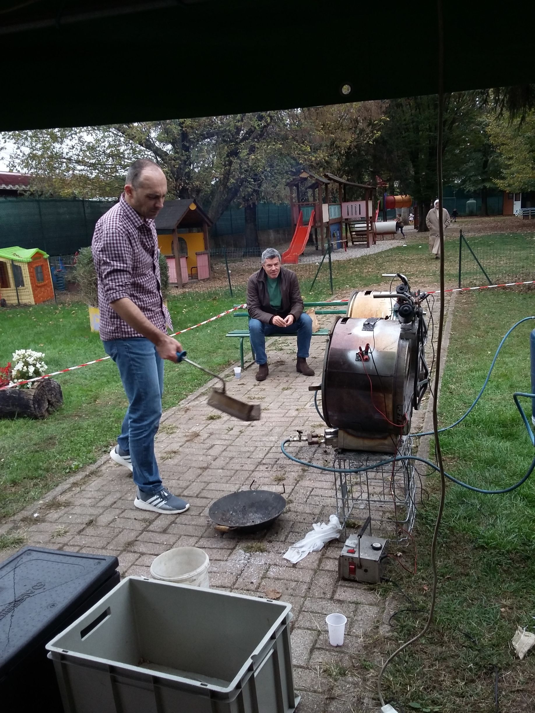 Un uomo si occupa di un barbecue affumicatore in un cortile, con un'altra persona seduta nelle vicinanze e un braciere davanti.