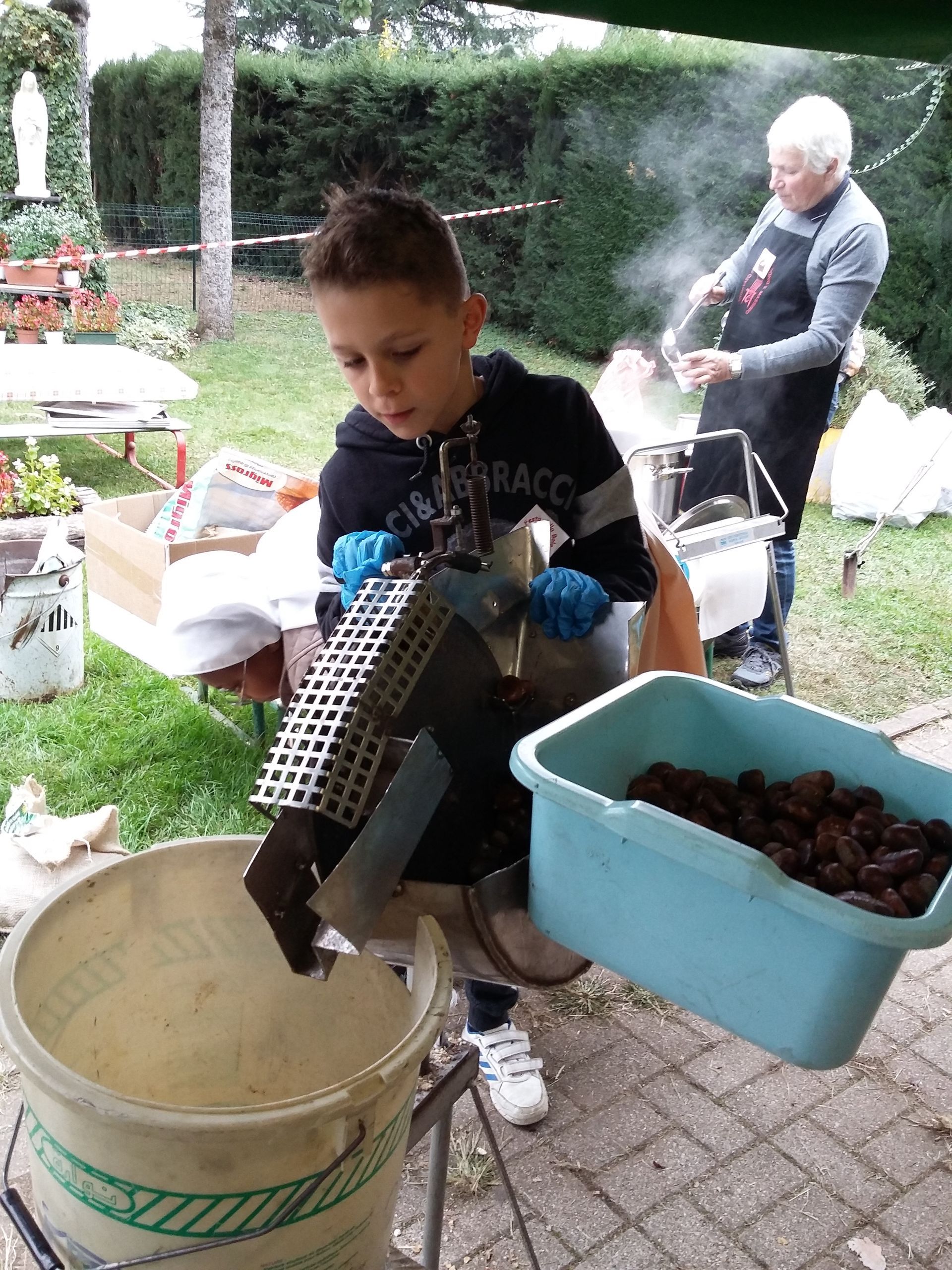 Un bambino all'aperto tiene in mano un grande secchio nero sopra una vasca beige, con un'altra persona sullo sfondo e attrezzi da giardino nelle vicinanze.
