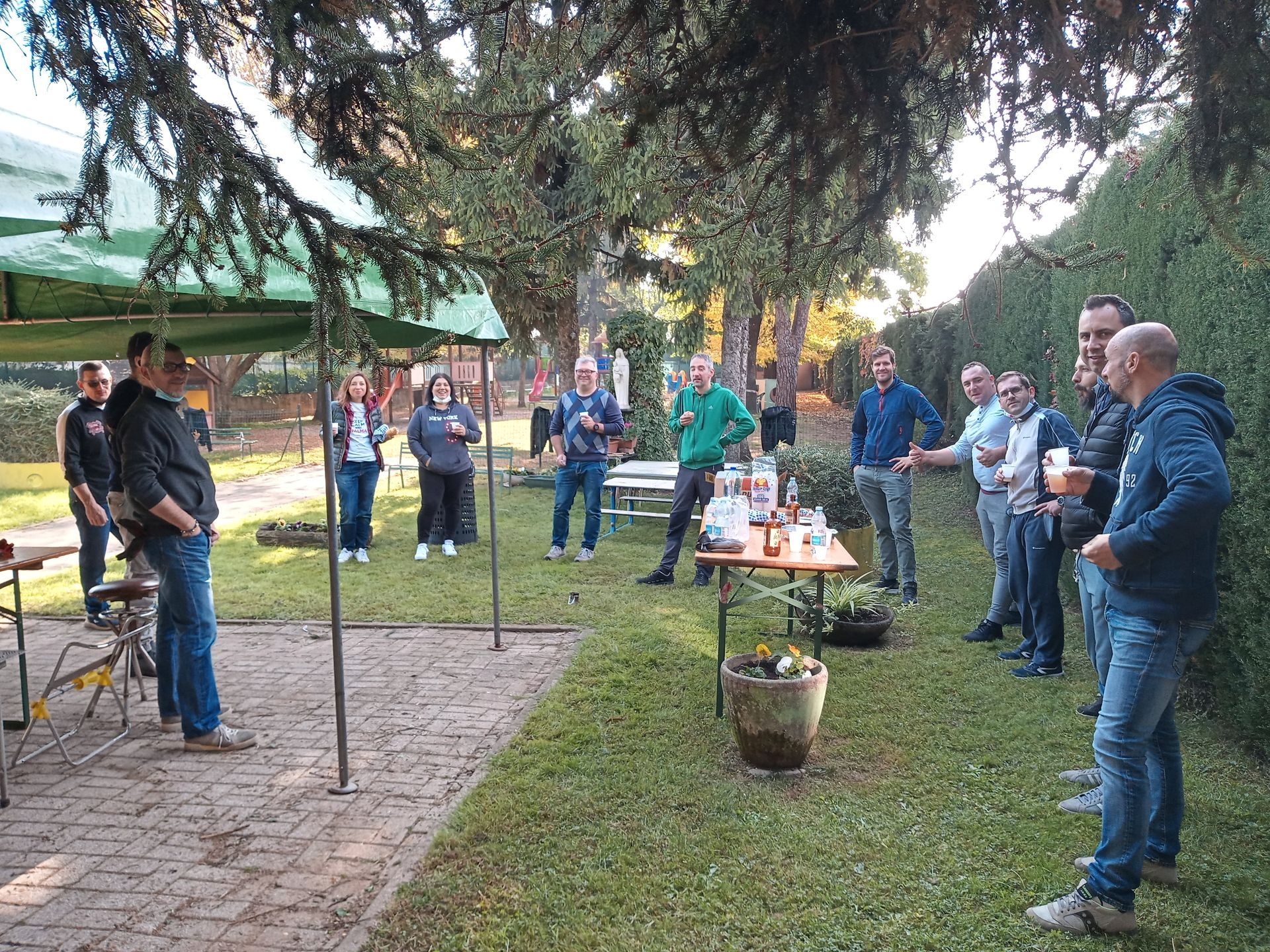 Un gruppo di persone si trova all'aperto in un parco, riunite attorno a un tavolino sotto gli alberi per un incontro informale.