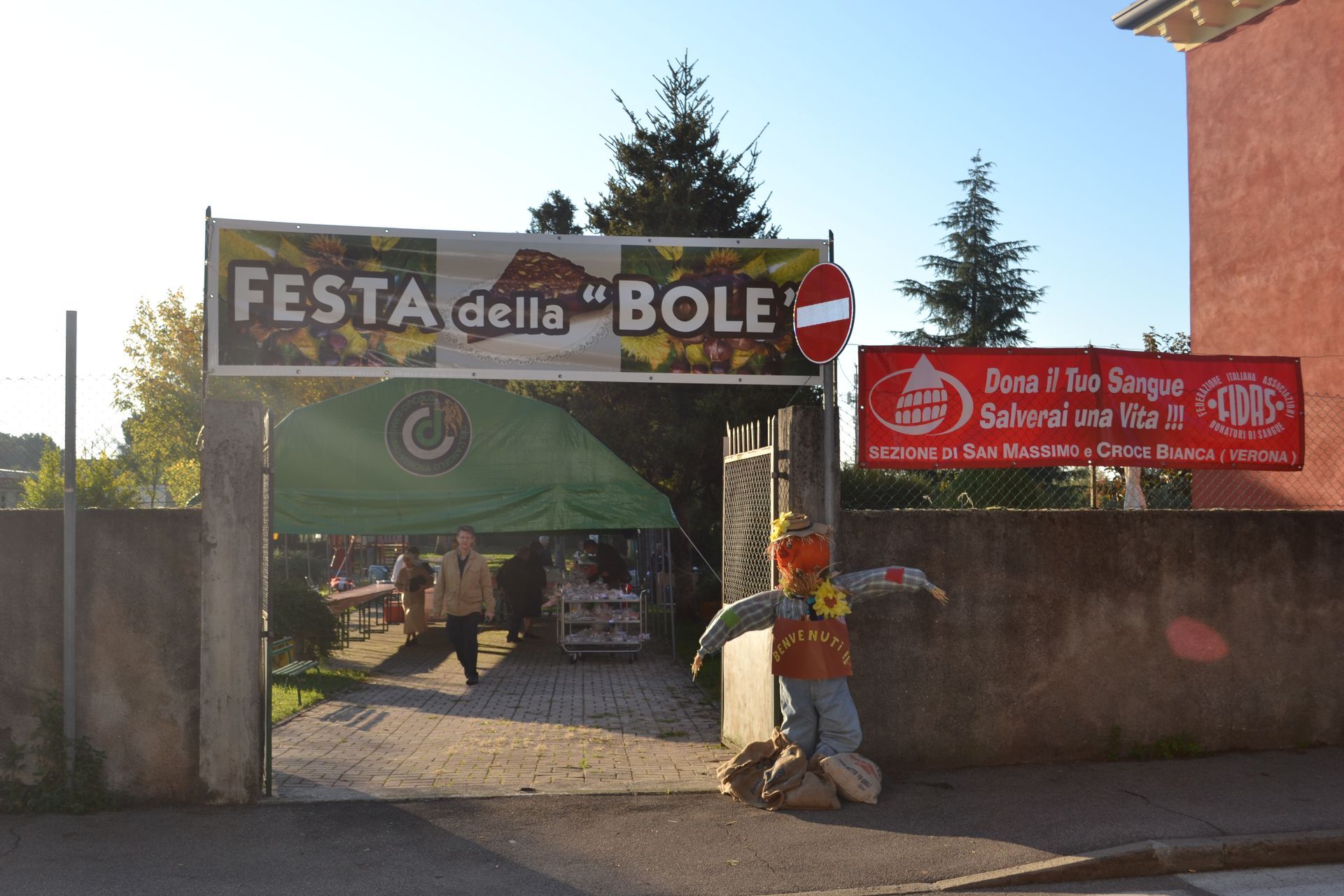 Ingresso alla Festa della Bole con striscioni colorati, una persona vicino al cancello e un cartello rosso sul muro