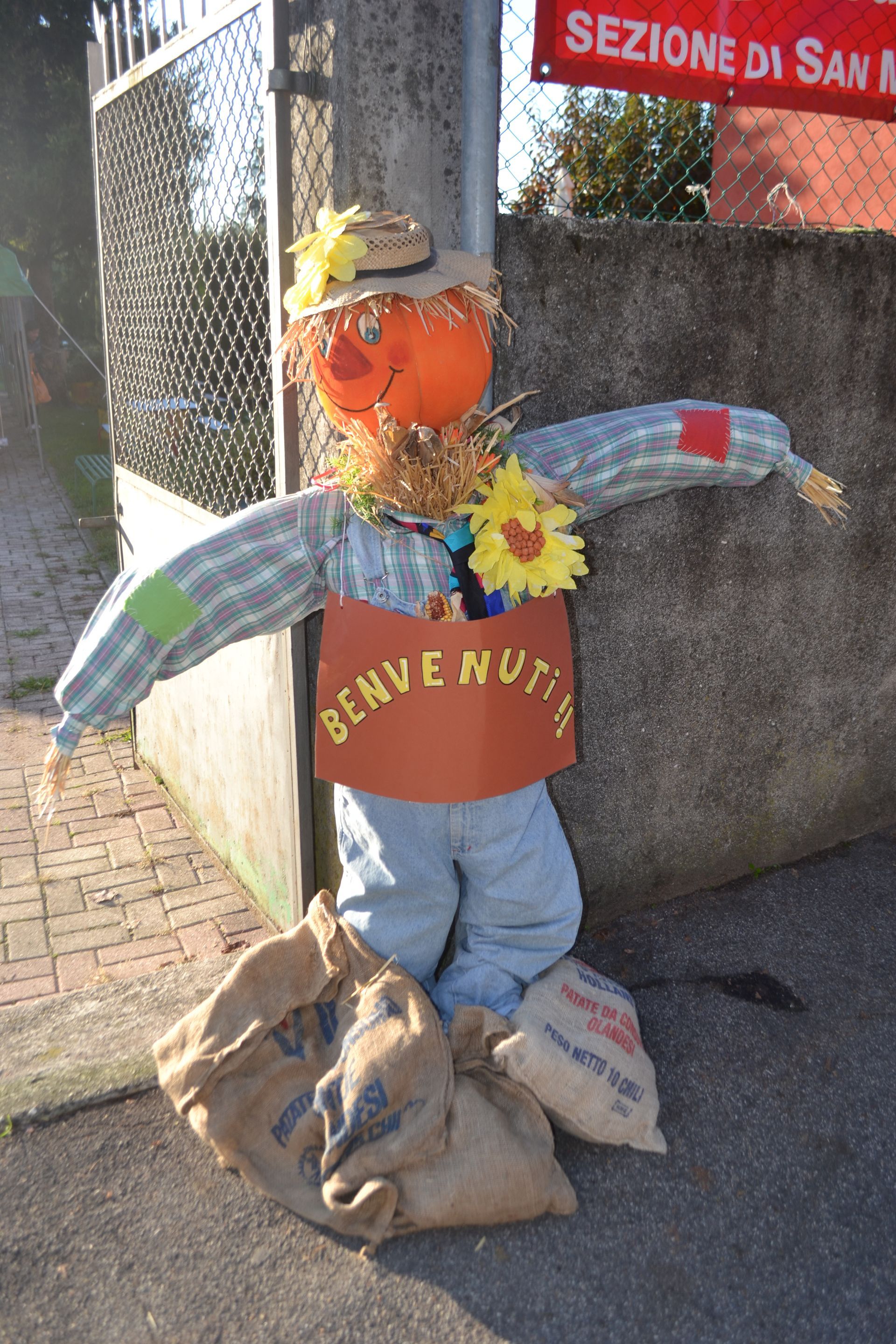 Spaventapasseri con testa di zucca arancione, faccia di girasole e braccia di paglia accanto a un cartello e sacchi all'aperto