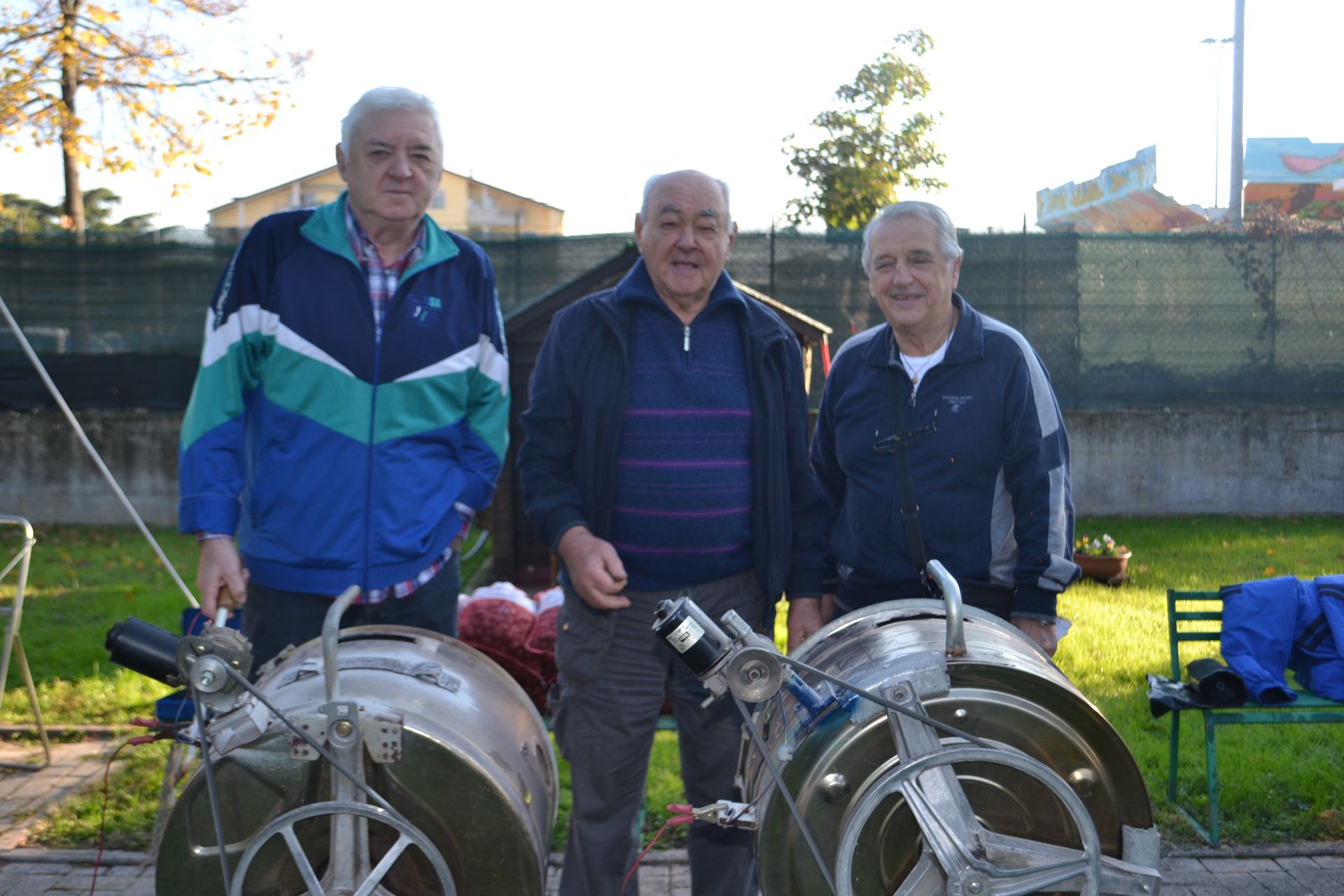 Tre persone in piedi all'aperto accanto a due ruote di metallo d'epoca in un campo soleggiato.
