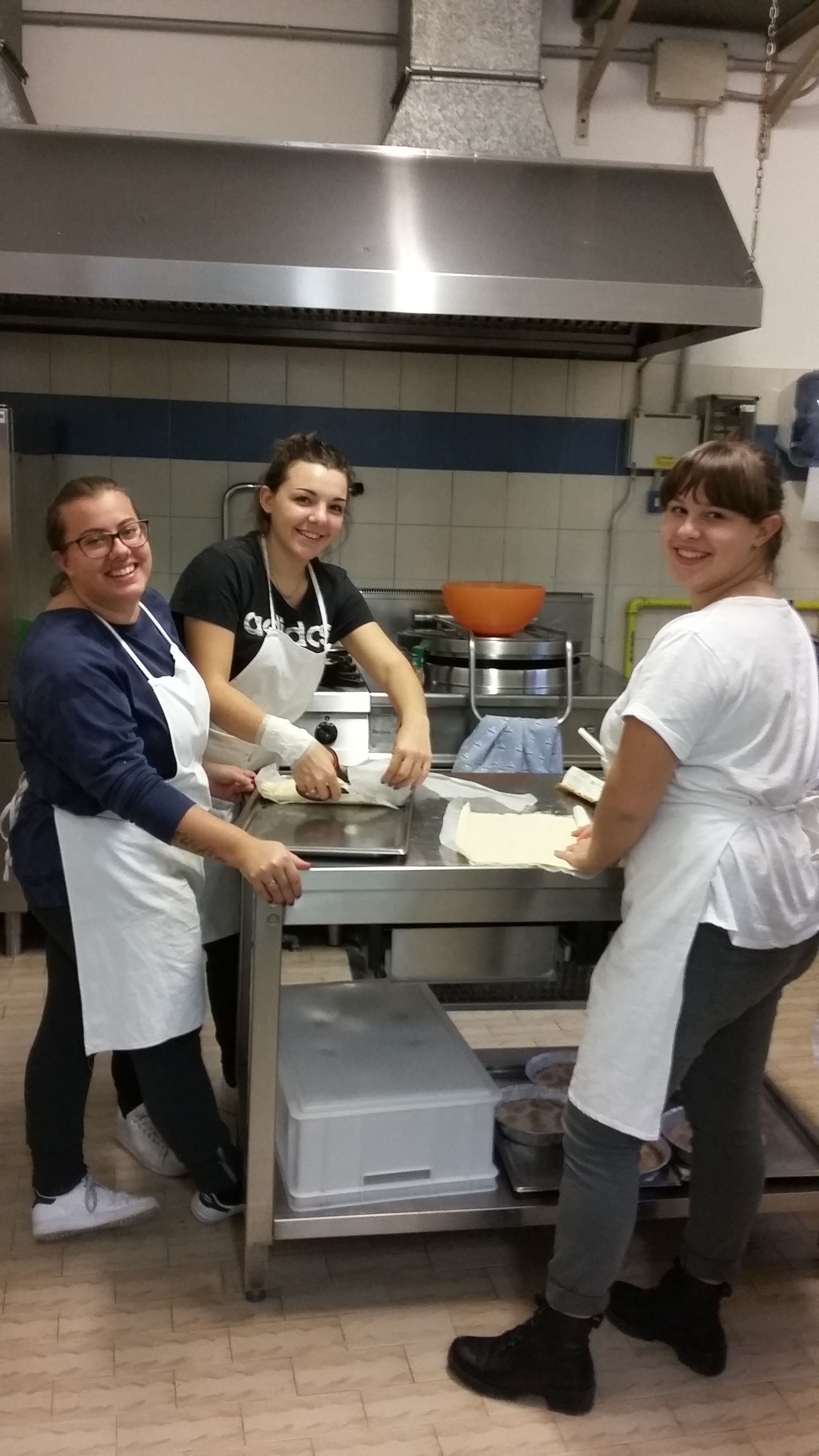 Tre persone con il grembiule sorridono mentre preparano del cibo in una cucina professionale.