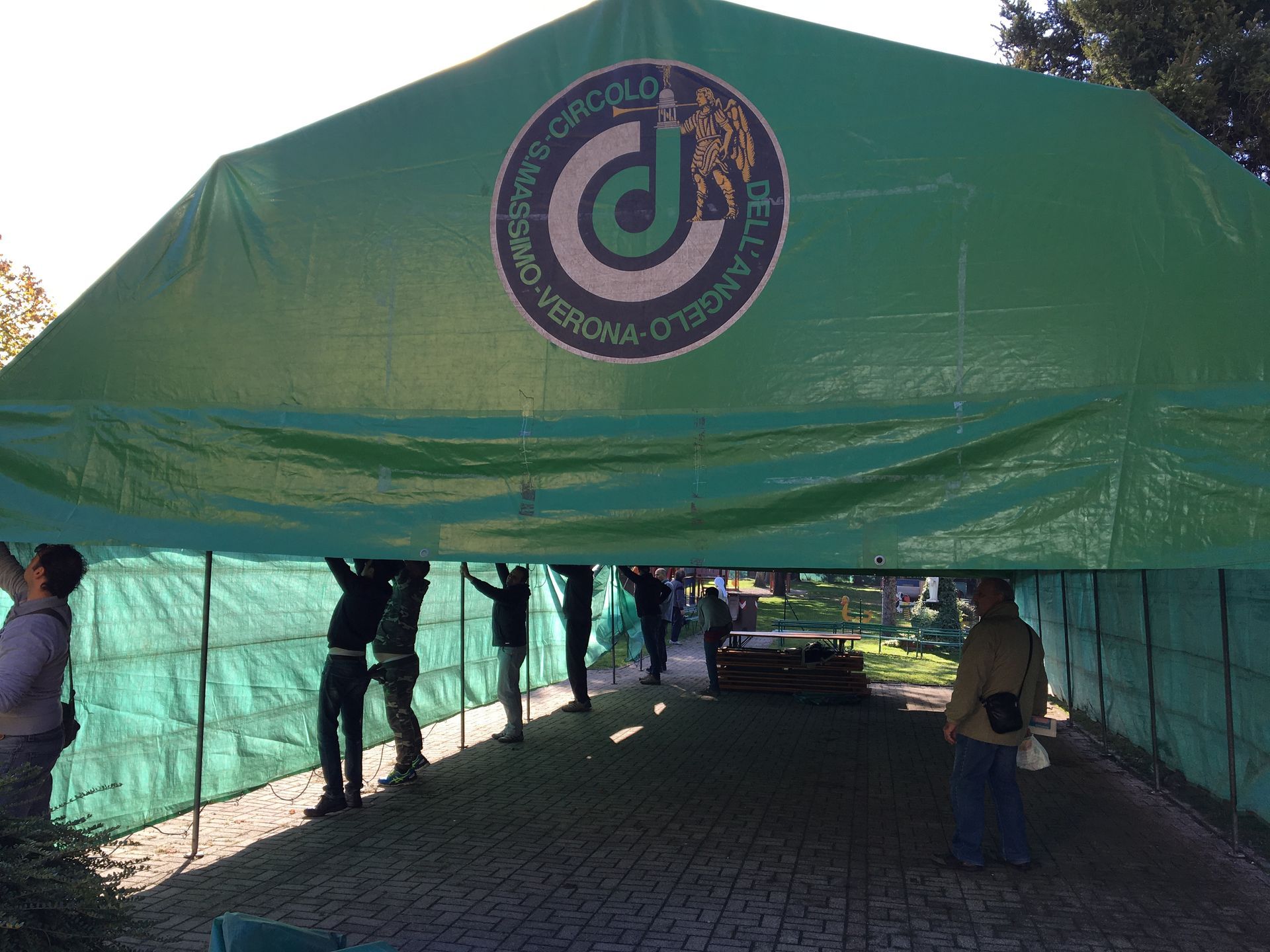 Una tettoia verde con un logo, che ripara le persone in piedi su un sentiero di ghiaia.
