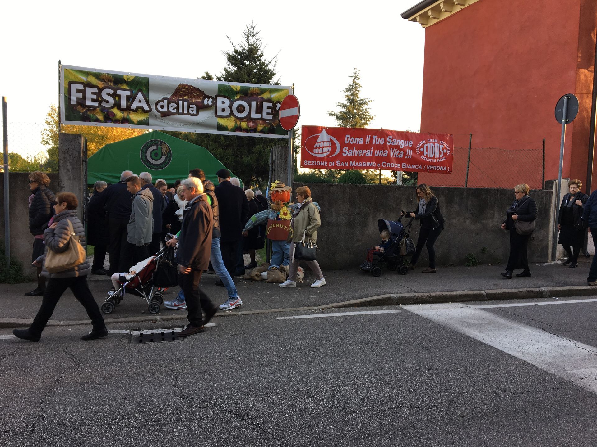 Persone che passano davanti a uno striscione festivo all'ingresso e a un cartello rosso all'esterno di un edificio all'angolo di una strada.