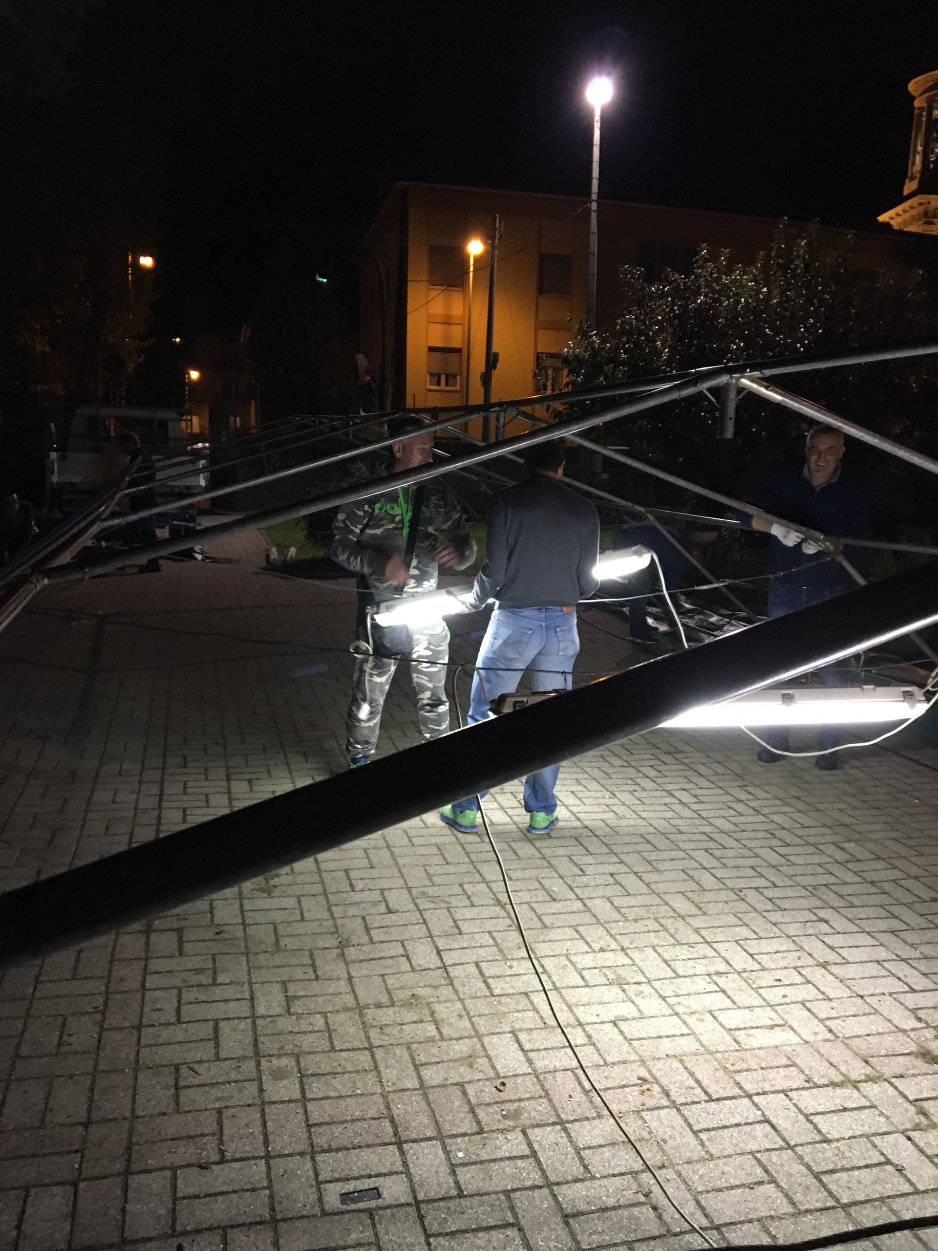 Due persone camminano di notte sotto una struttura metallica crollata in un cortile pavimentato illuminato.