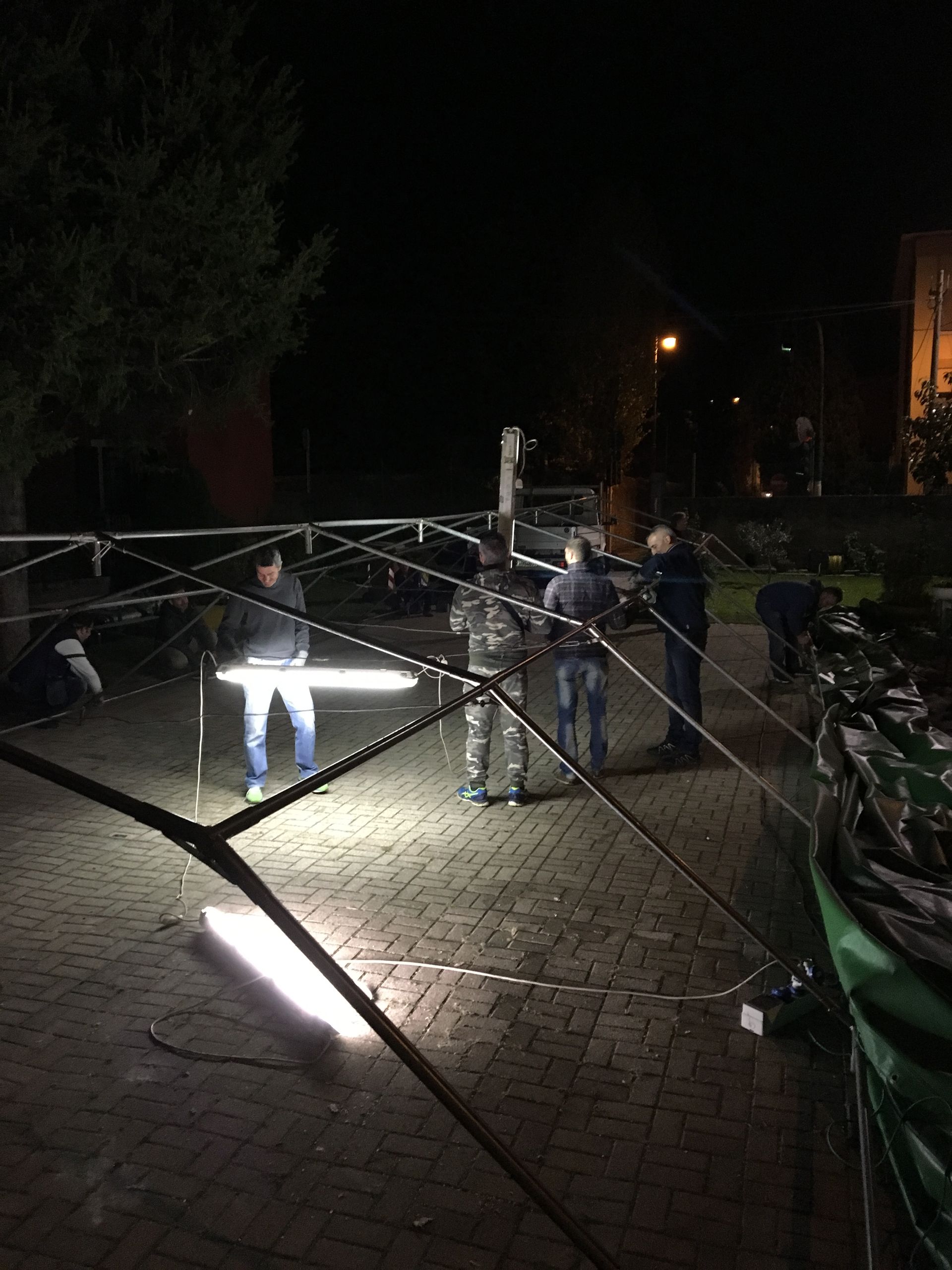 Di notte, in un cortile buio, le persone si radunavano attorno a una luce intensa, con delle biciclette parcheggiate nelle vicinanze.