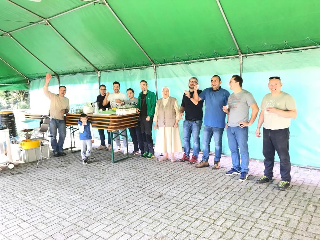 Gruppo di persone in posa sotto una tettoia verde accanto a pile di assi di legno su un pavimento pavimentato.