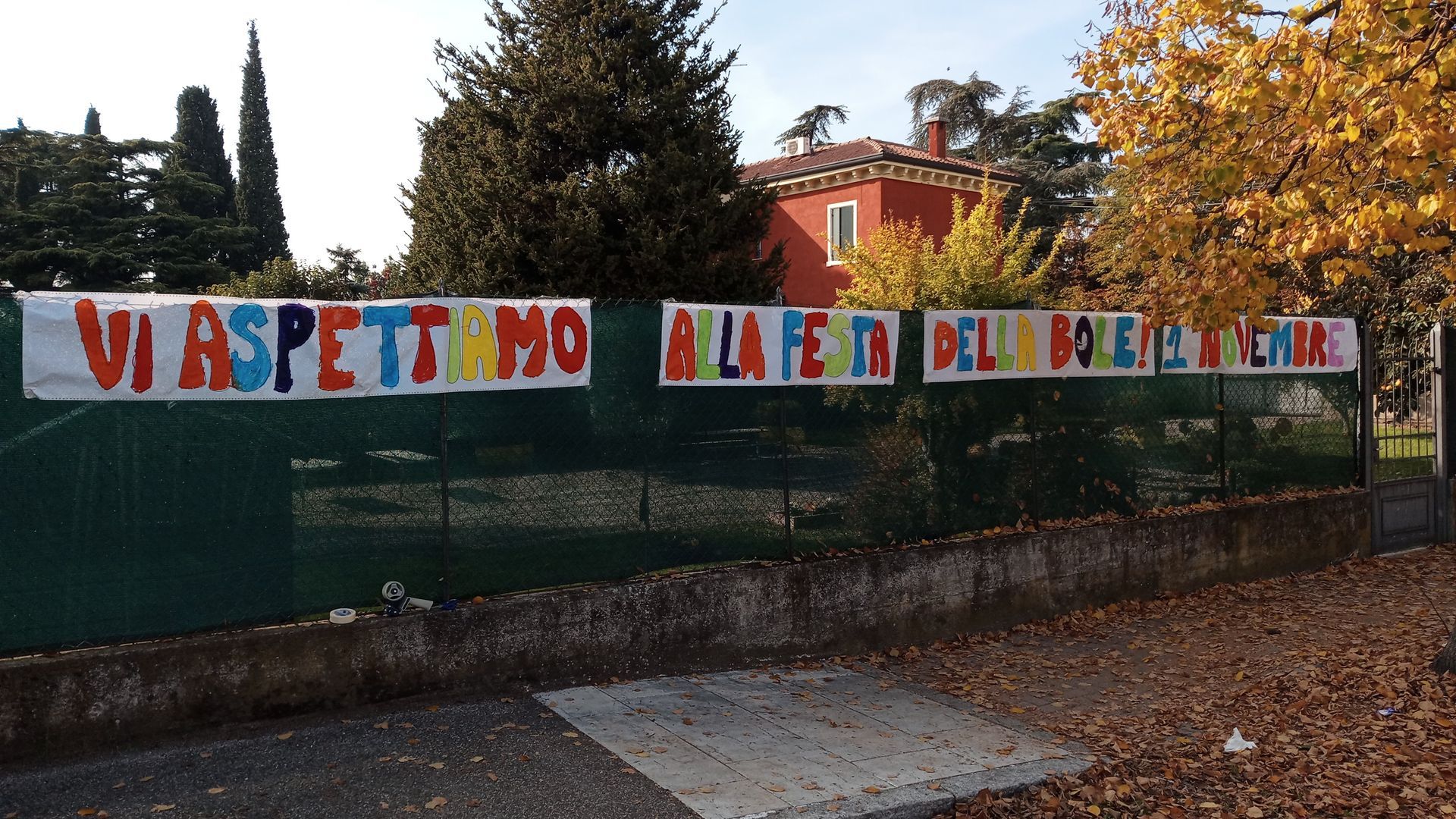 Uno striscione di protesta colorato appeso a una recinzione in un quartiere alberato, con case e alberi sullo sfondo.