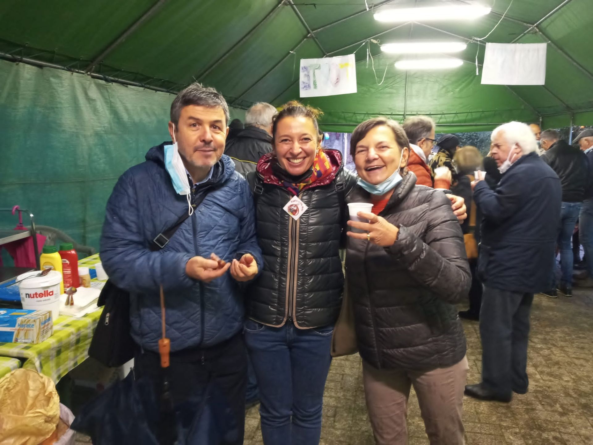 Tre persone sorridenti all'interno di una bancarella del mercato, con in mano delle tazze, sotto una tenda verde.