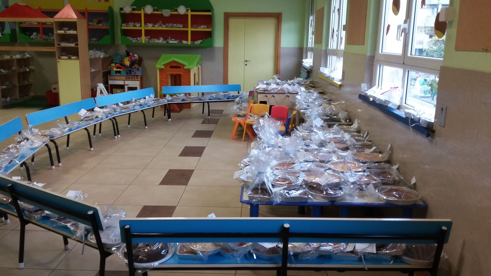 In una luminosa mensa scolastica con finestre e panche, lunghi tavoli apparecchiati con pasti confezionati.
