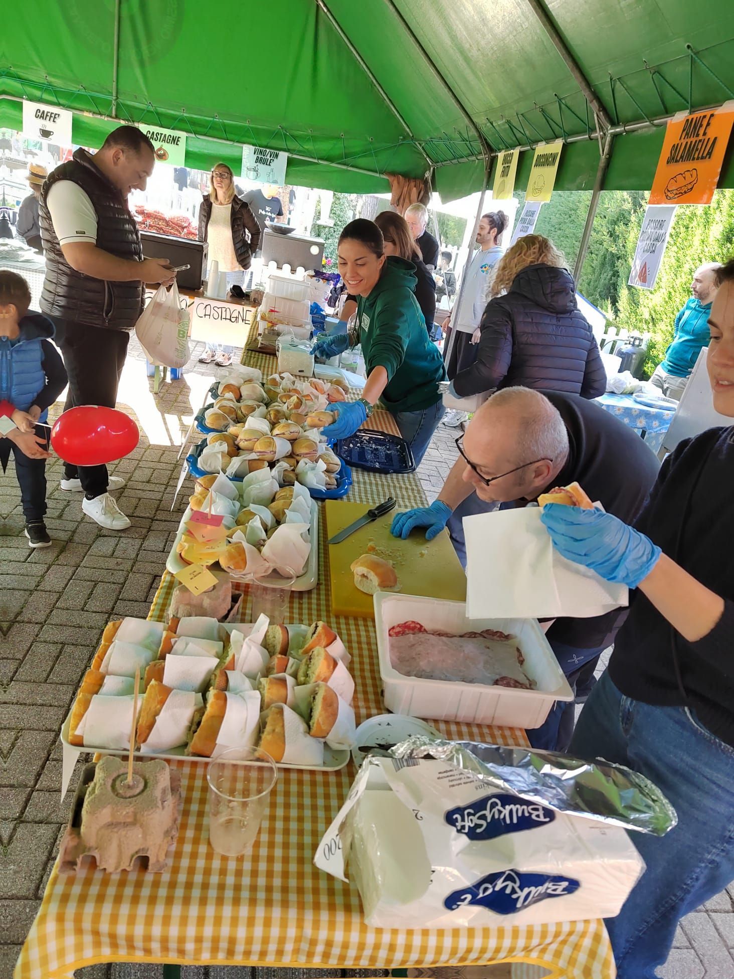 Bancarella di cibo sotto un tendone verde con venditori che offrono snack e bevande confezionati a un tavolo a scacchi.