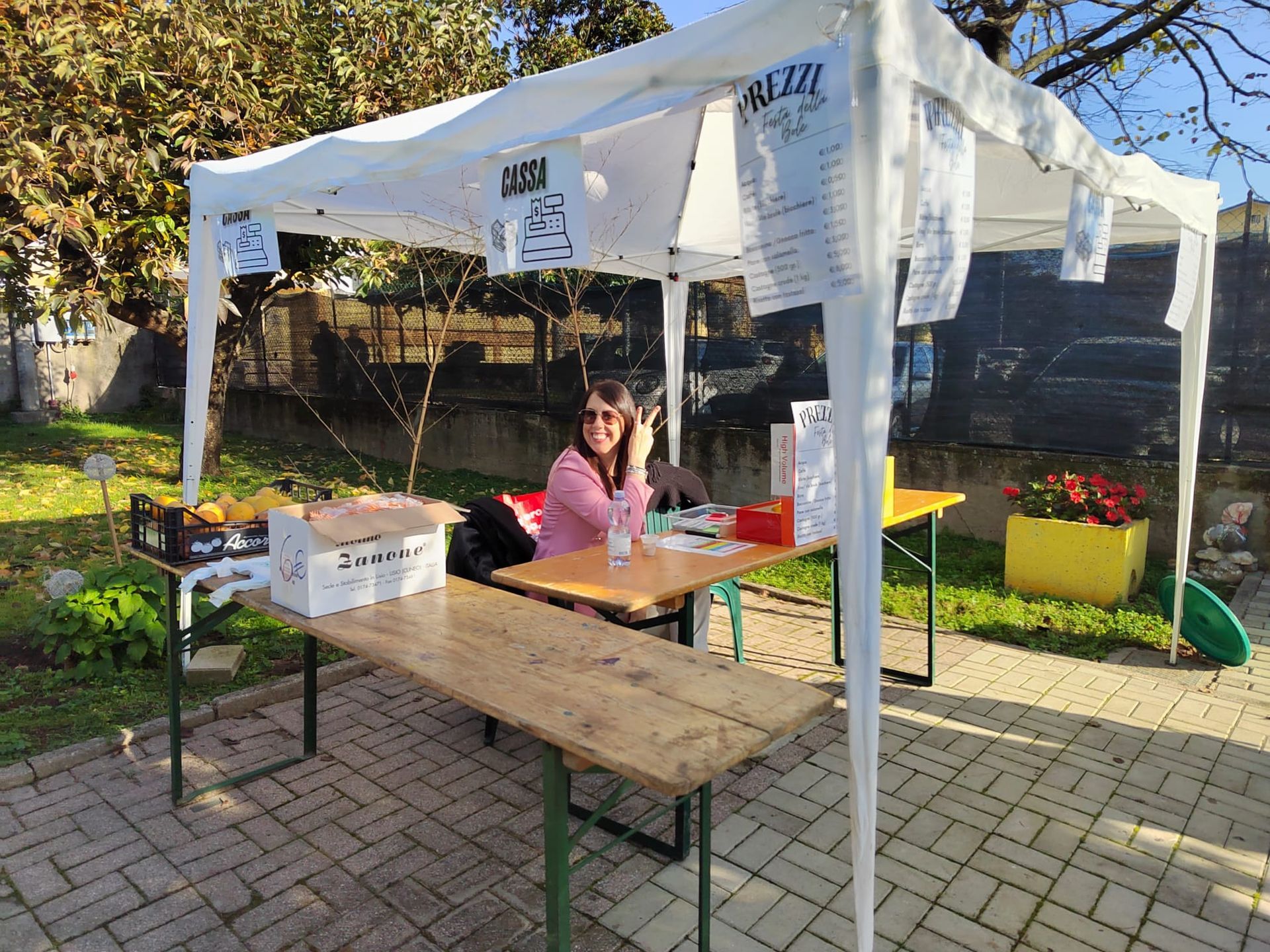Persona seduta a un tavolo di legno sotto una tenda bianca in un mercato all'aperto soleggiato.