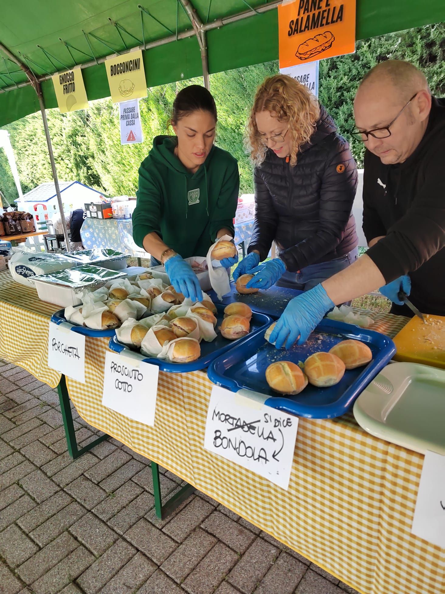 Tre persone con i guanti servono panini in una bancarella di un mercato all'aperto sotto una tenda verde.