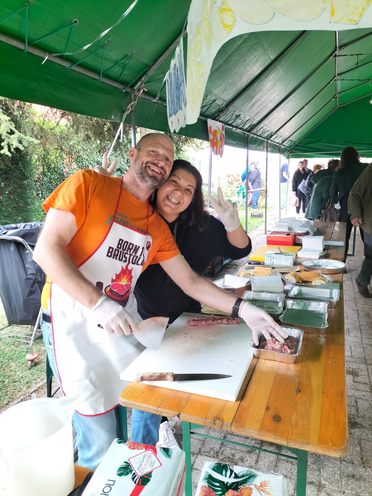 Due persone sorridenti preparano del cibo a un tavolo sotto una tenda verde.