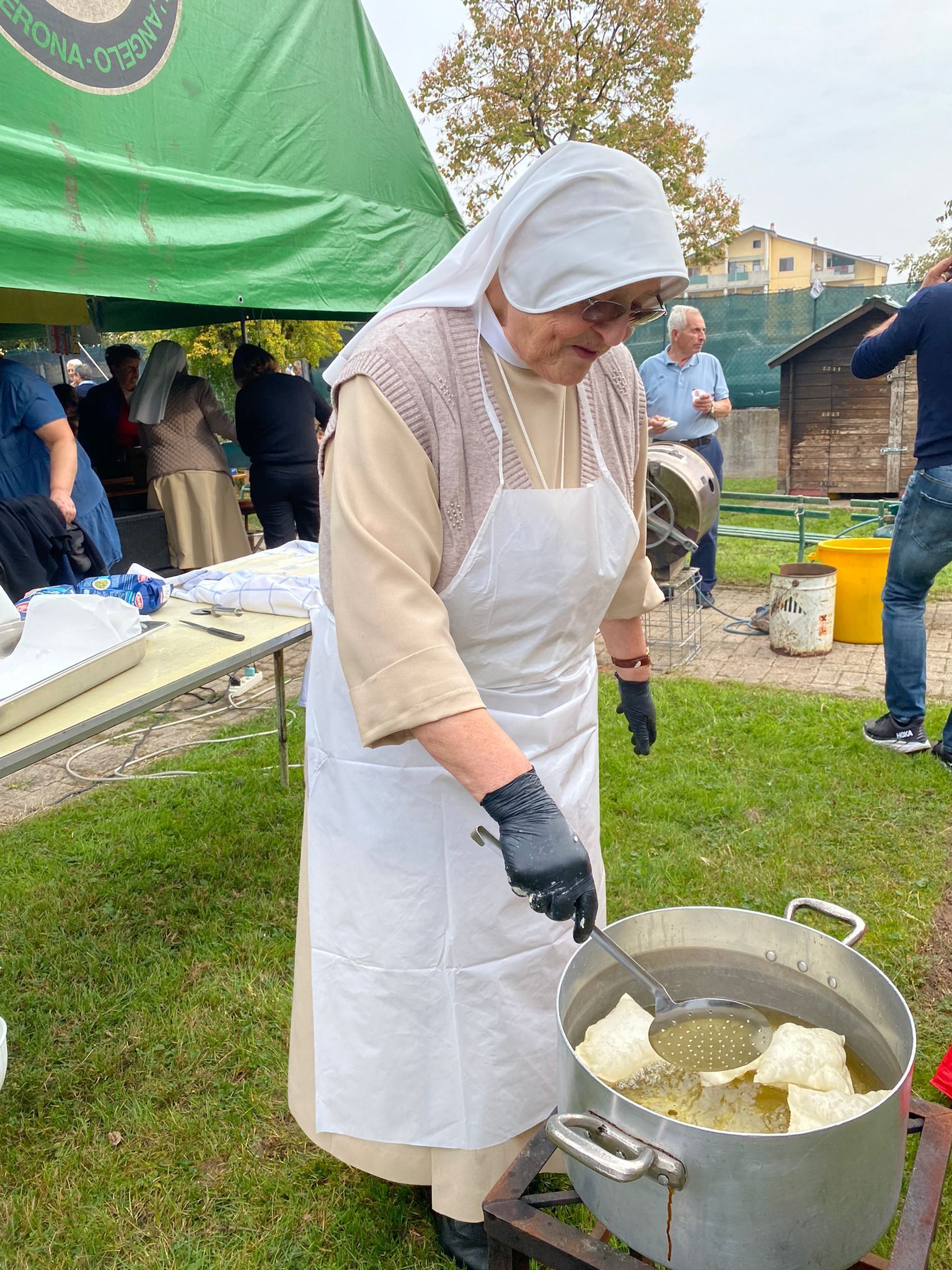 Una persona con grembiule e cappuccio bianchi mescola qualcosa in una grande pentola all'aperto durante un evento culinario.