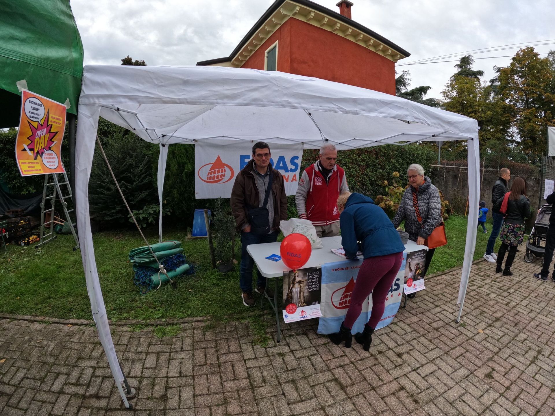 Persone che allestiscono uno stand sotto un gazebo bianco su un sentiero lastricato, con palloncini e striscioni.
