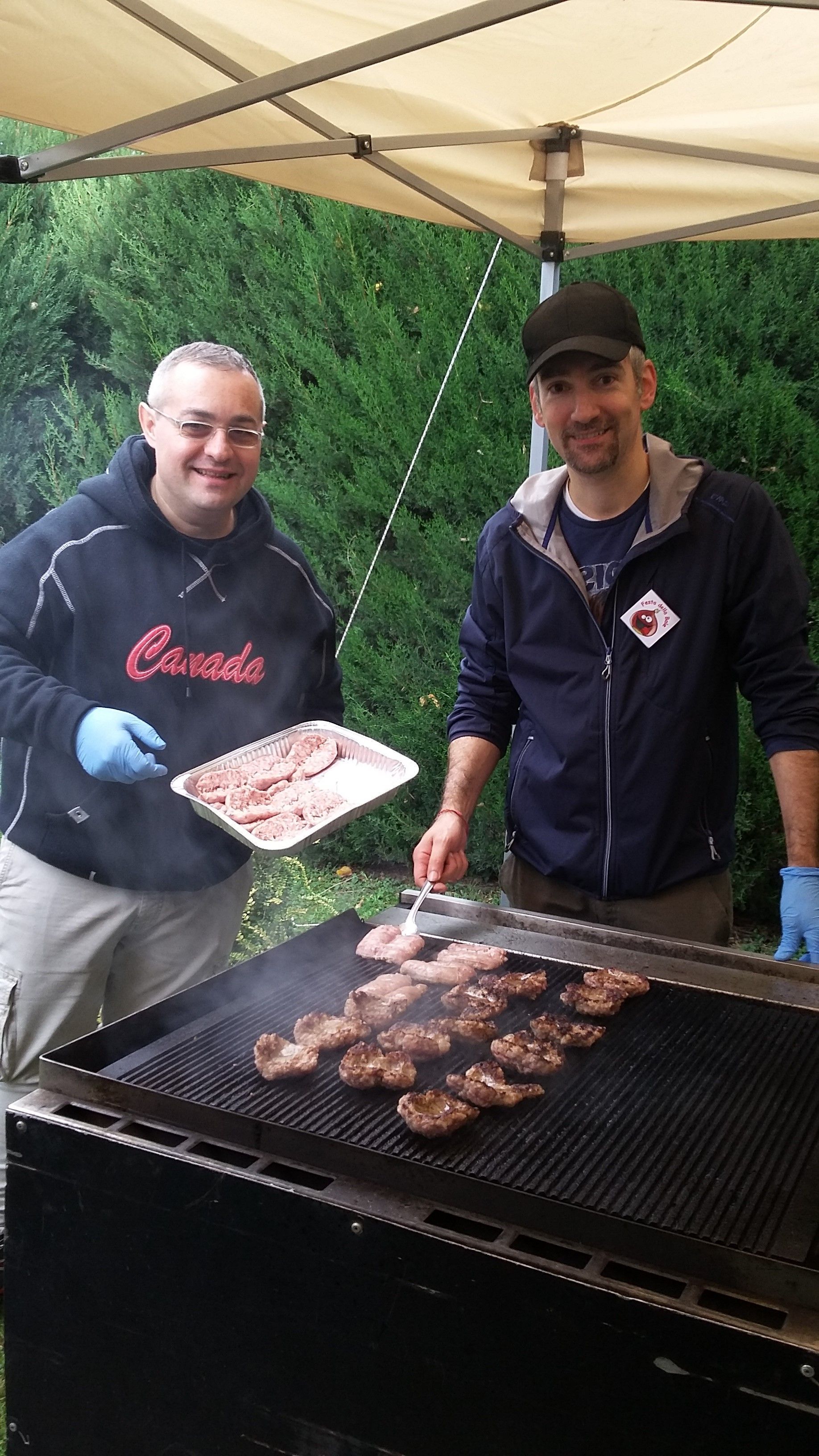 Due persone grigliano carne sotto una tettoia, una tiene un vassoio e l'altra si occupa del barbecue.
