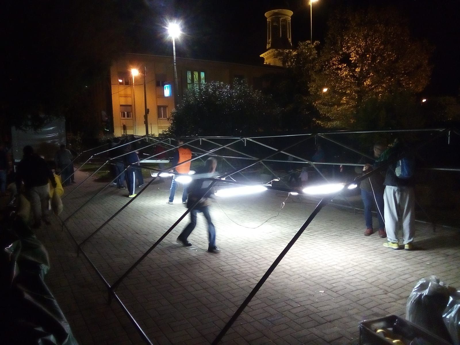 Piazza notturna con persone accanto a una struttura circolare illuminata e luci brillanti a terra.
