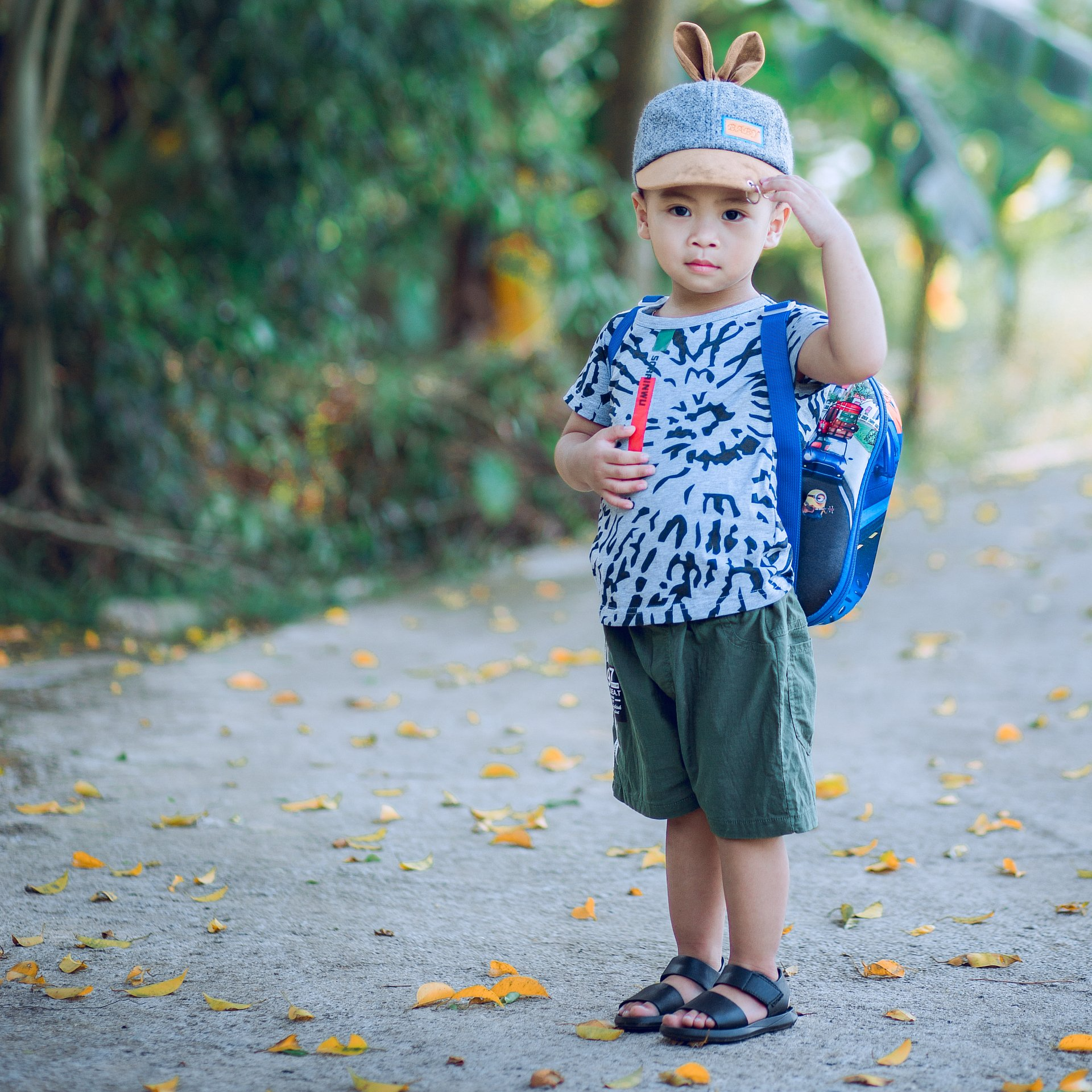 Bambino in piedi su un sentiero che tiene in mano un giocattolo, indossa un berretto blu, una maglietta a fantasia, pantaloncini verdi e uno zainetto.