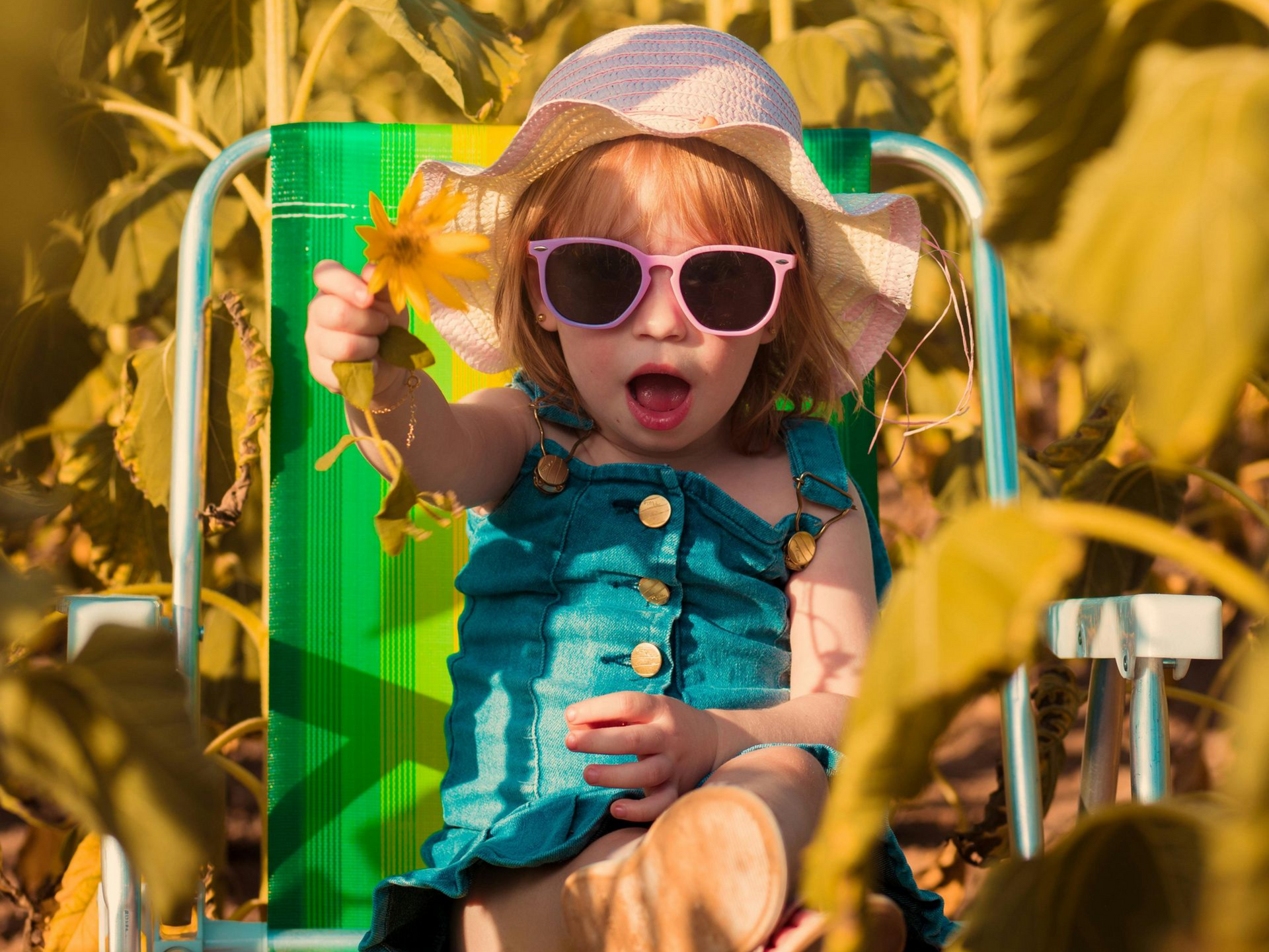Un bambino piccolo con occhiali da sole e cappello rosa è seduto su una sedia verde, tenendo in mano un fiore giallo in un campo soleggiato.
