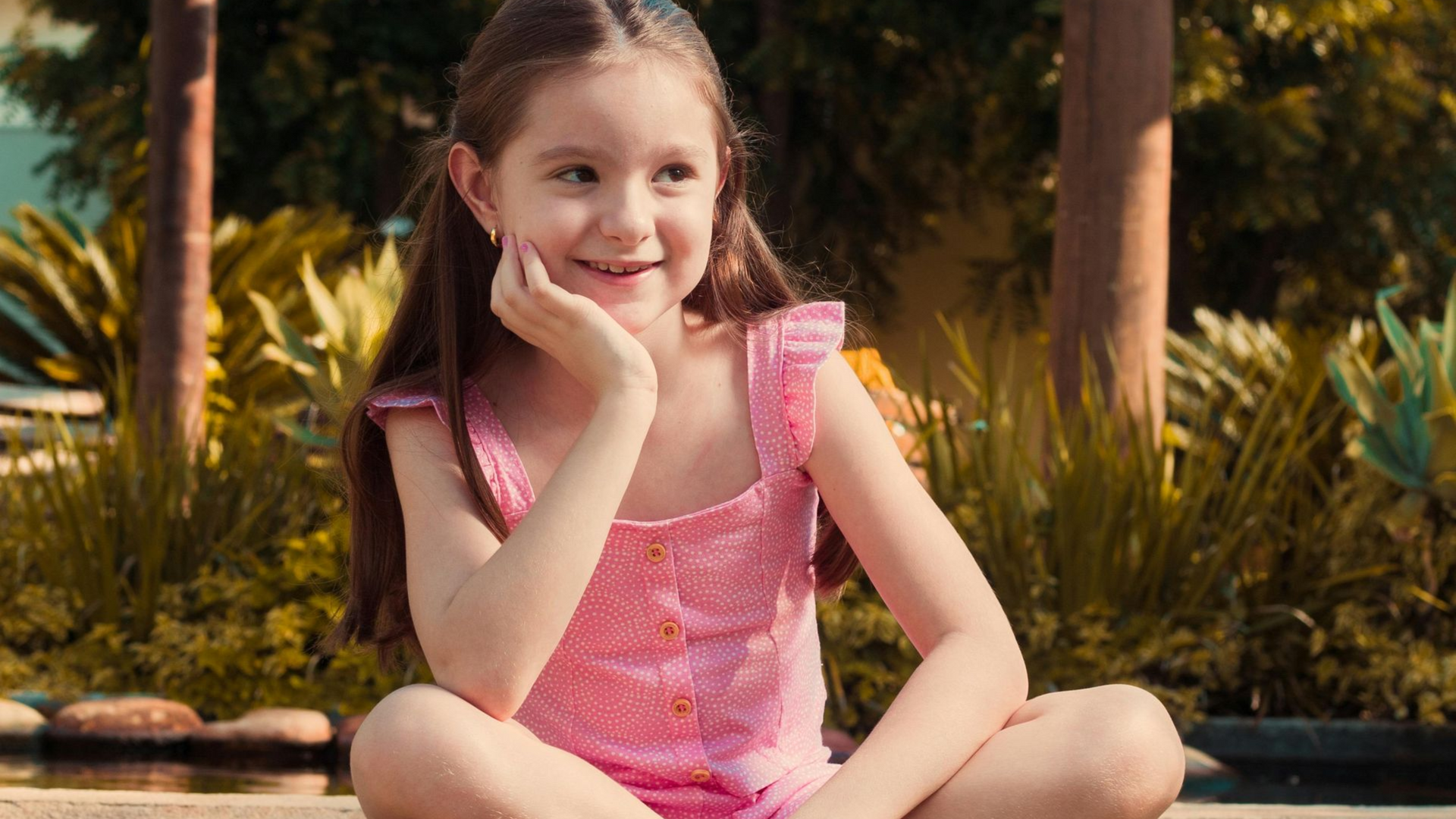 Bambina sorridente in costume da bagno rosa seduta a bordo piscina con piante sullo sfondo.