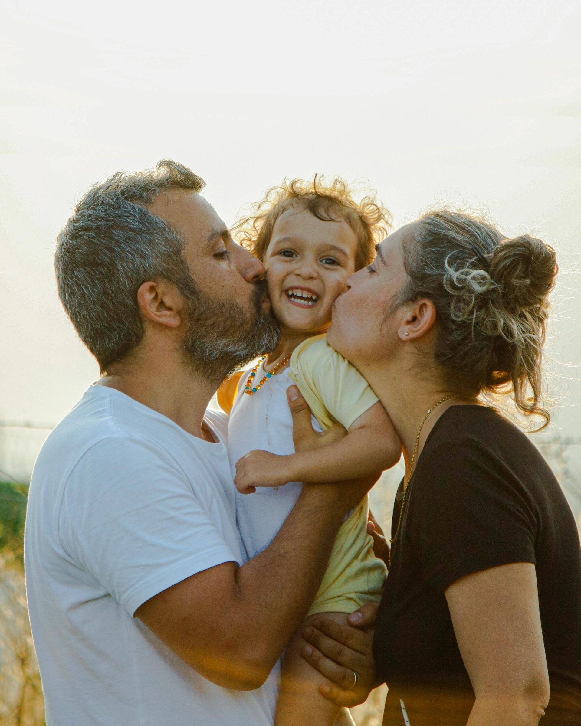 Una famiglia si abbraccia all'aperto al tramonto, un bambino sorridente tra due adulti che gli baciano entrambe le guance.