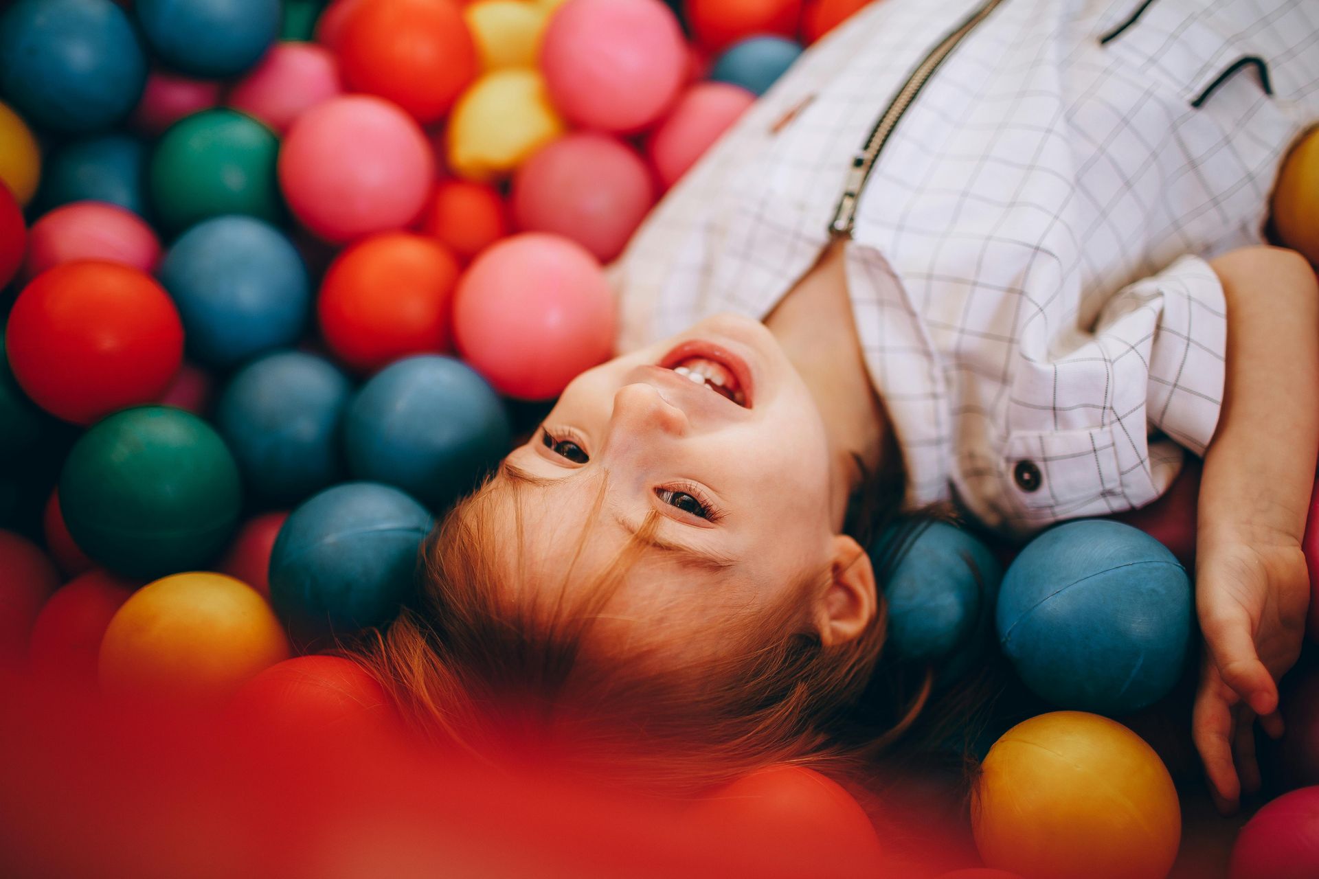 Bambino sdraiato in una piscina di palline colorate, sorride alla telecamera.