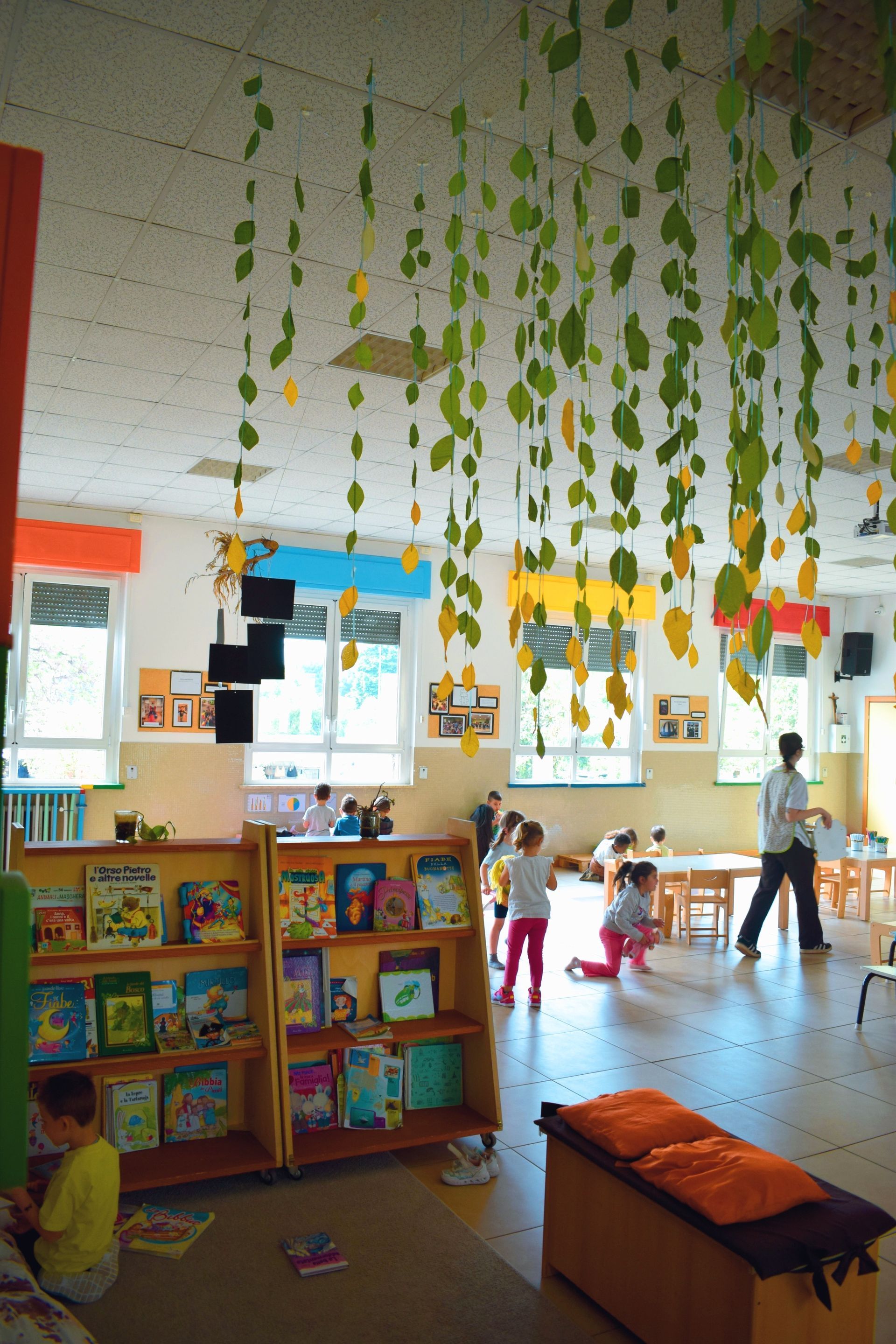 Aula scolastica con foglie di carta appese, scaffali pieni di libri e bambini che giocano vicino a grandi finestre.