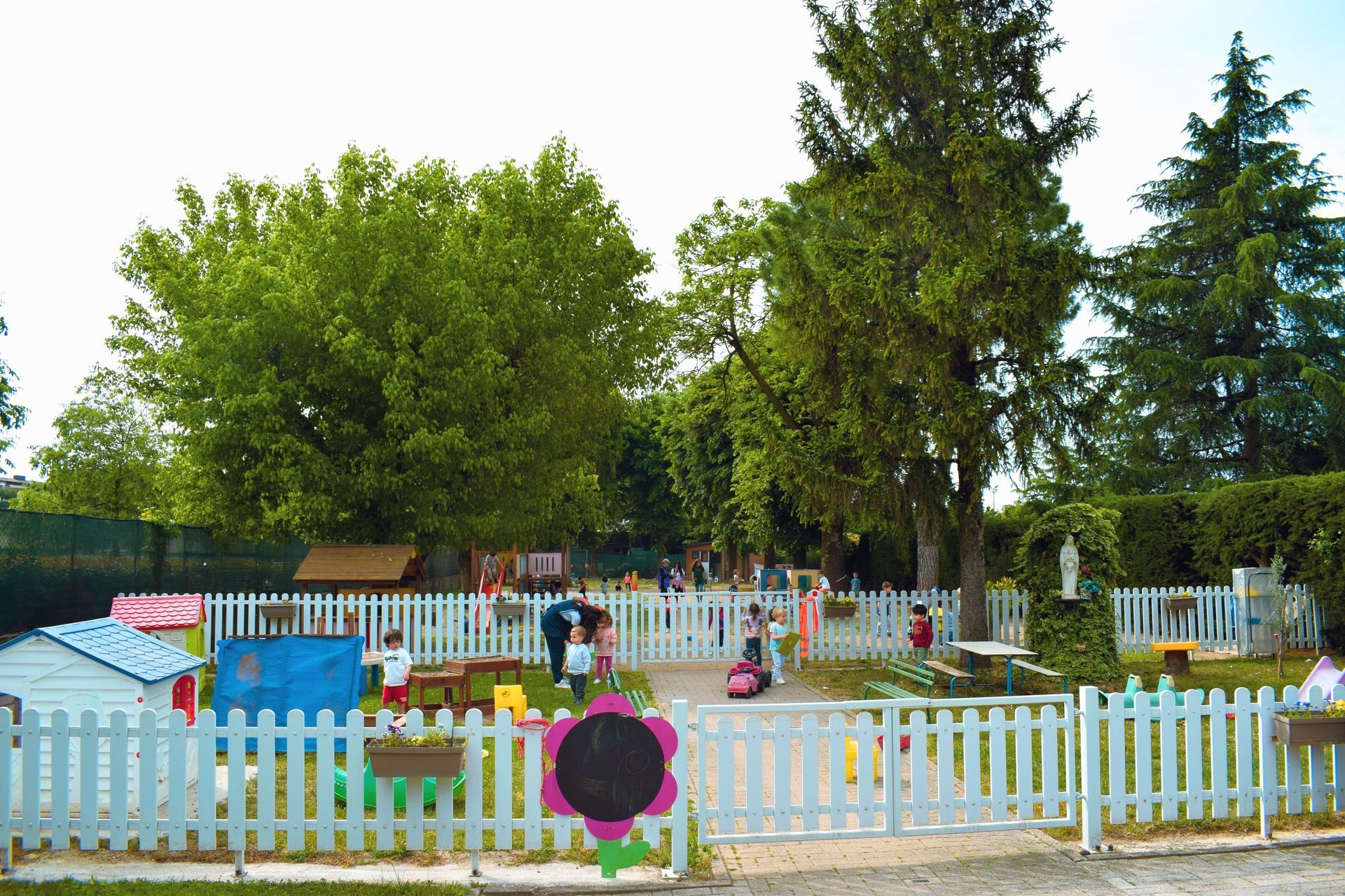 Parco giochi colorato con una staccionata bianca, attrezzature blu e alti alberi sullo sfondo.
