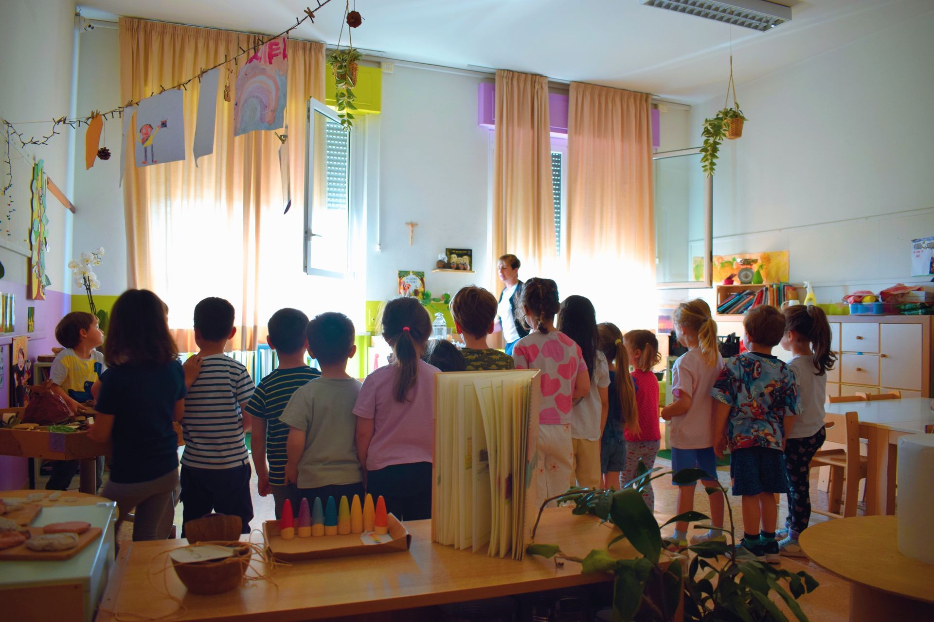 Bambini in piedi in un'aula decorata, che guardano una presentazione davanti alla classe, con tende e decorazioni appese.