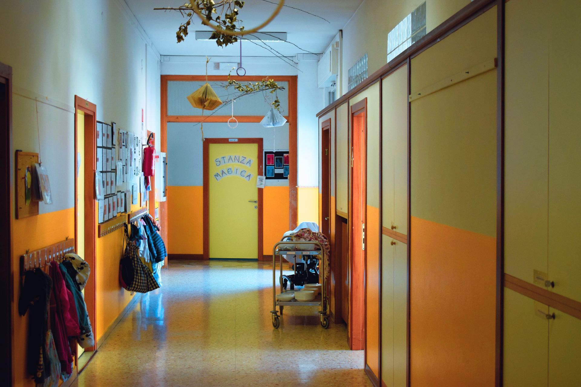 Corridoio scolastico vuoto con pareti arancioni e gialle, armadietti e porte che conducono a una stanza illuminata di blu.