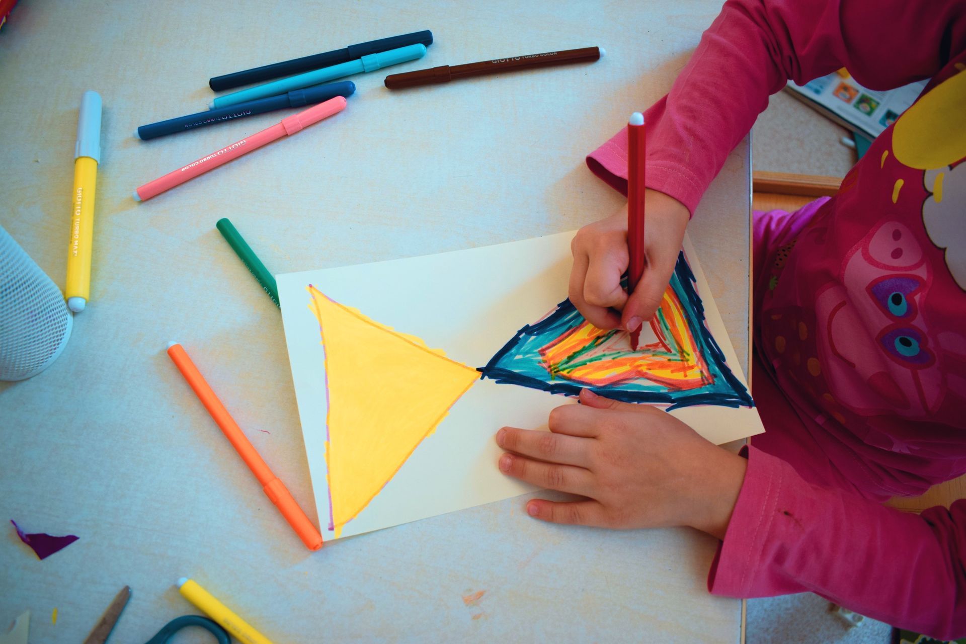 Un bambino disegna un triangolo colorato con i pennarelli su un foglio di carta seduto a un tavolo.