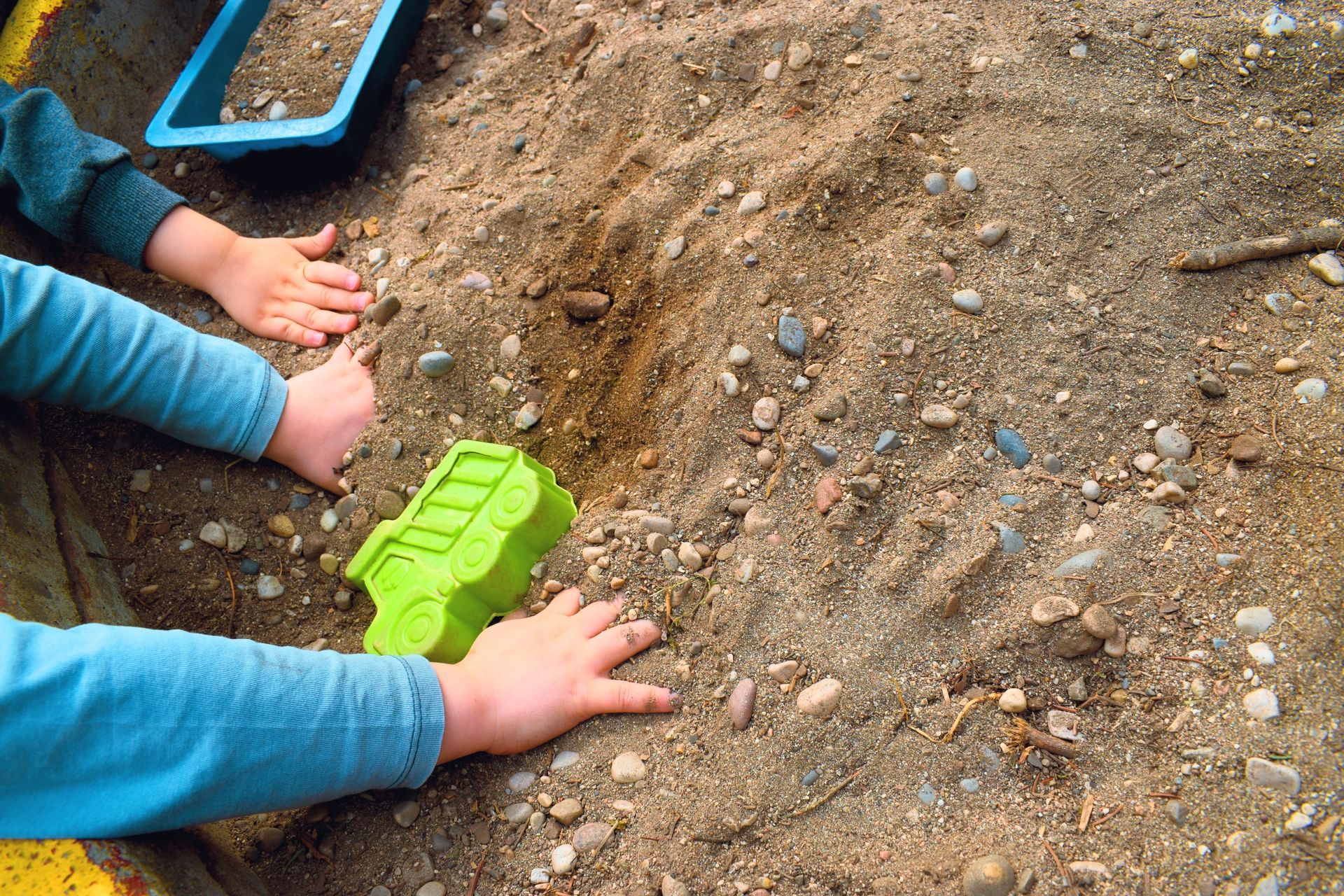 Due mani di bambini giocano nella terra con un camioncino giocattolo verde e una paletta blu.