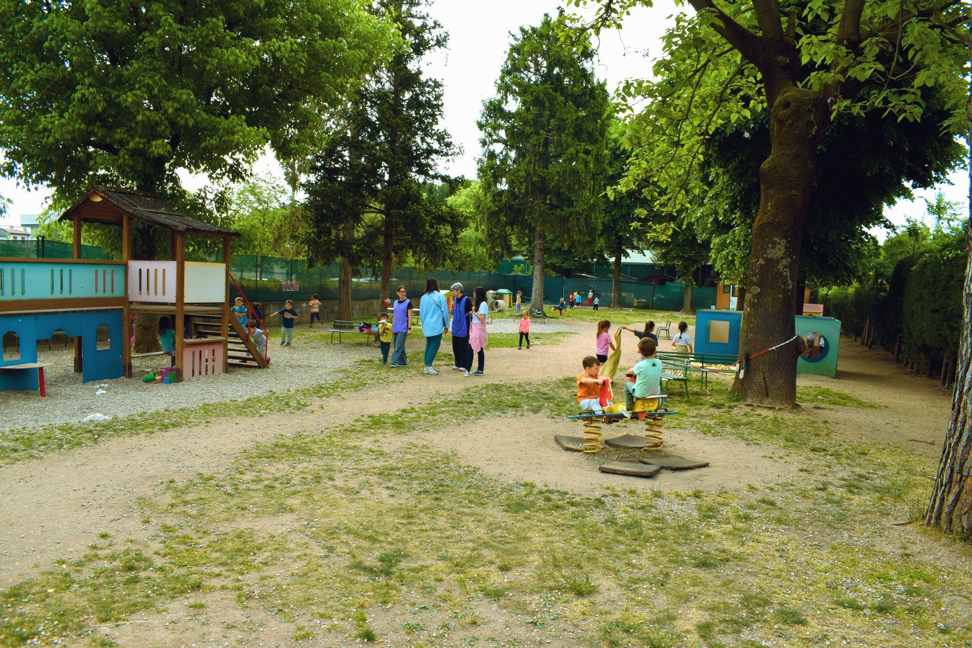 Persone in piedi in un parco erboso con giochi per bambini e alberi.