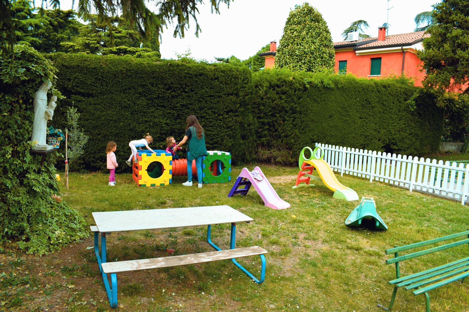 Bambini che giocano su colorate attrezzature da parco giochi in un prato con siepi e una recinzione bianca.
