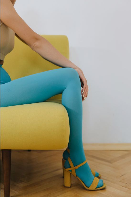 Vrouw zit op een gele stoel, draagt ​​een blauwe panty en gele hakken, haar arm rust op haar knie.