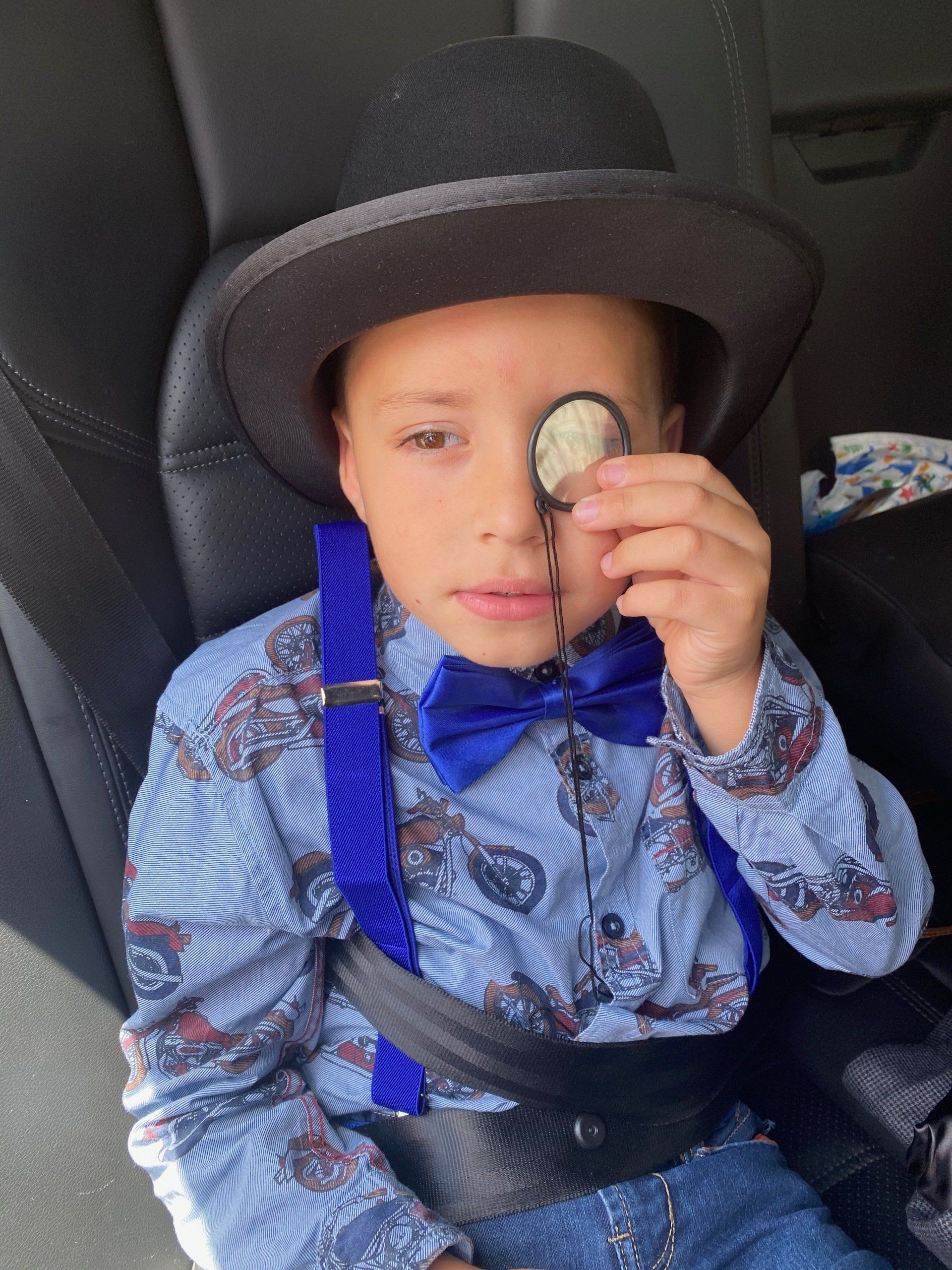 A young boy wearing a hat and bow tie is sitting in a car seat holding a magnifying glass.