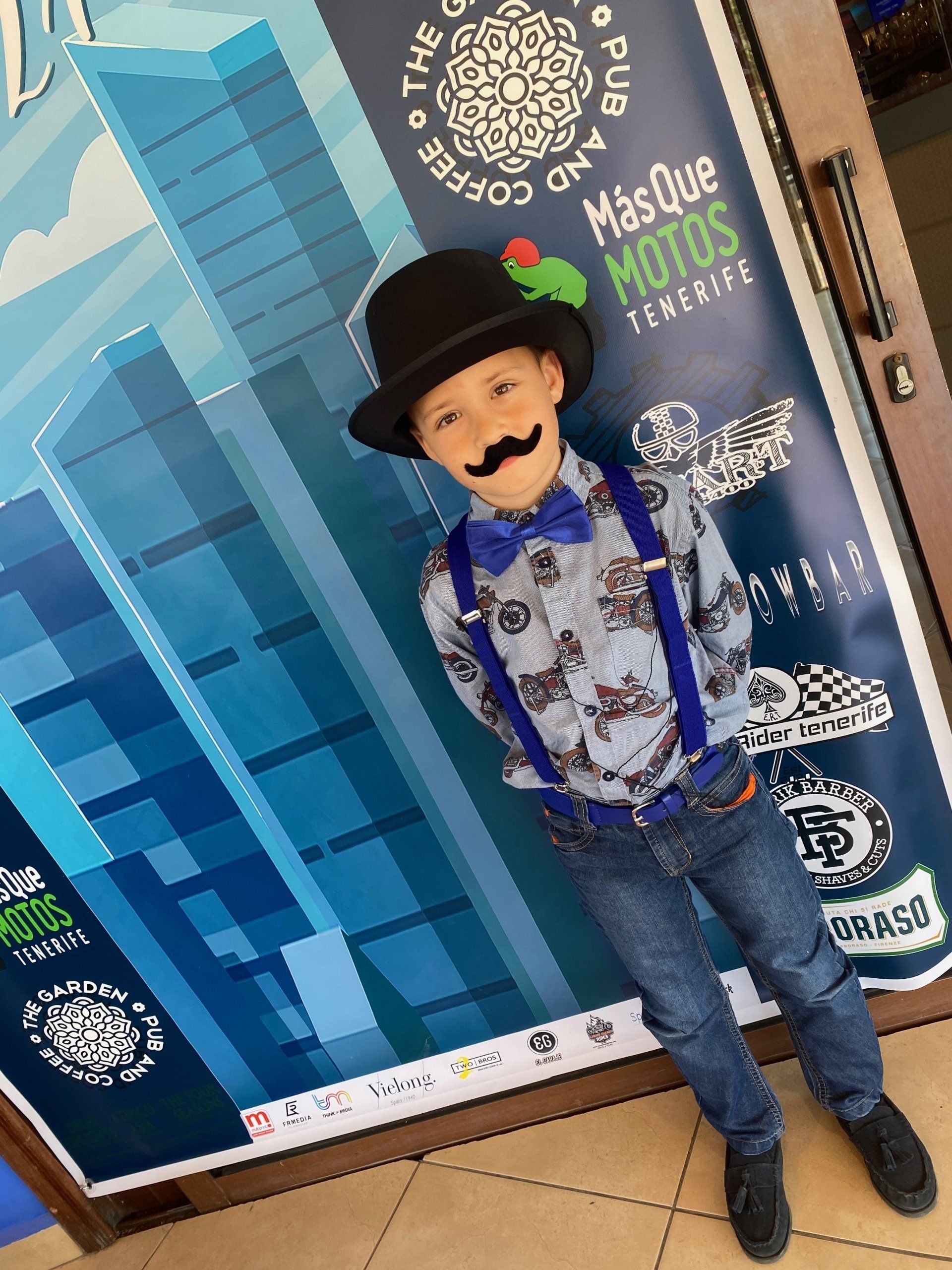 A young boy wearing a hat and suspenders is standing in front of a poster that says mes que motos
