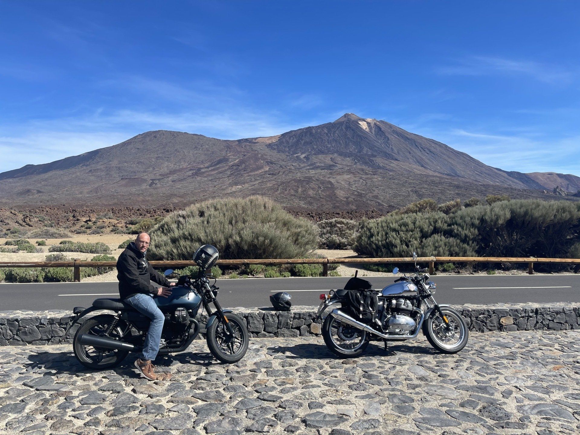 Two motorcycles are parked on the side of the road with a mountain in the background.