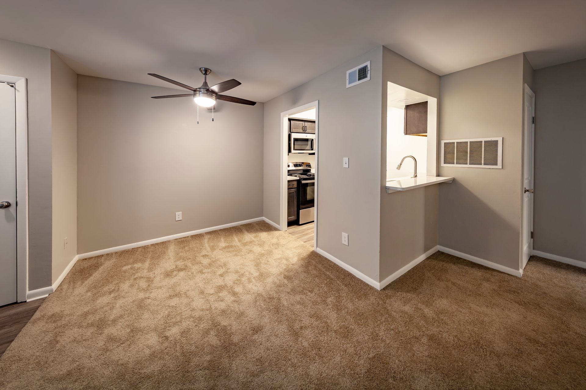 An empty living room with a ceiling fan and a kitchen in the background.